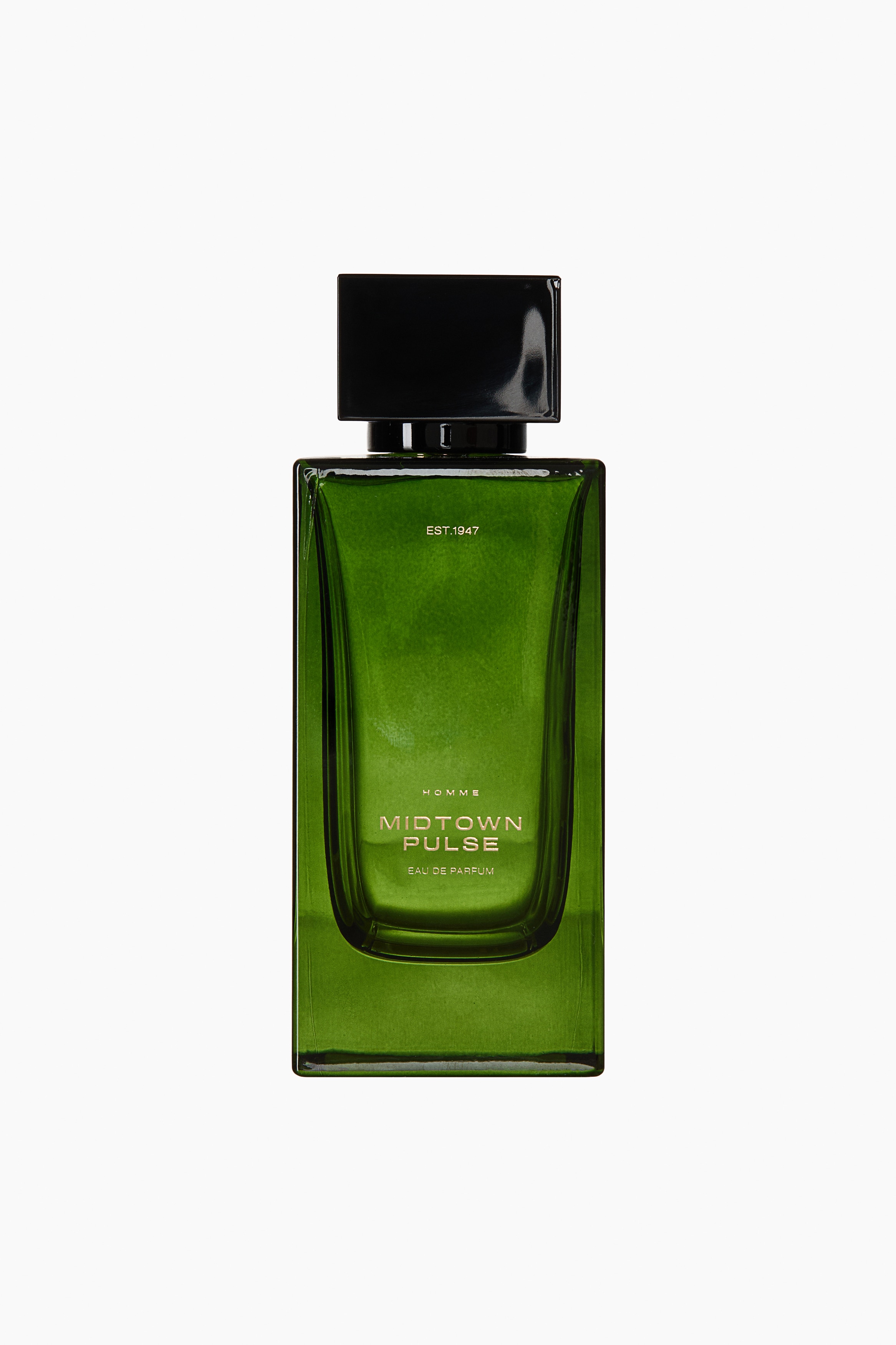 A rectangular green glass perfume bottle labeled "MIDTOWN PULSE" is standing upright in a straight-on view, topped with a square black cap. A slender piece of dark wood is leaning against its right side, with two small, dark tonka beans resting at its base.