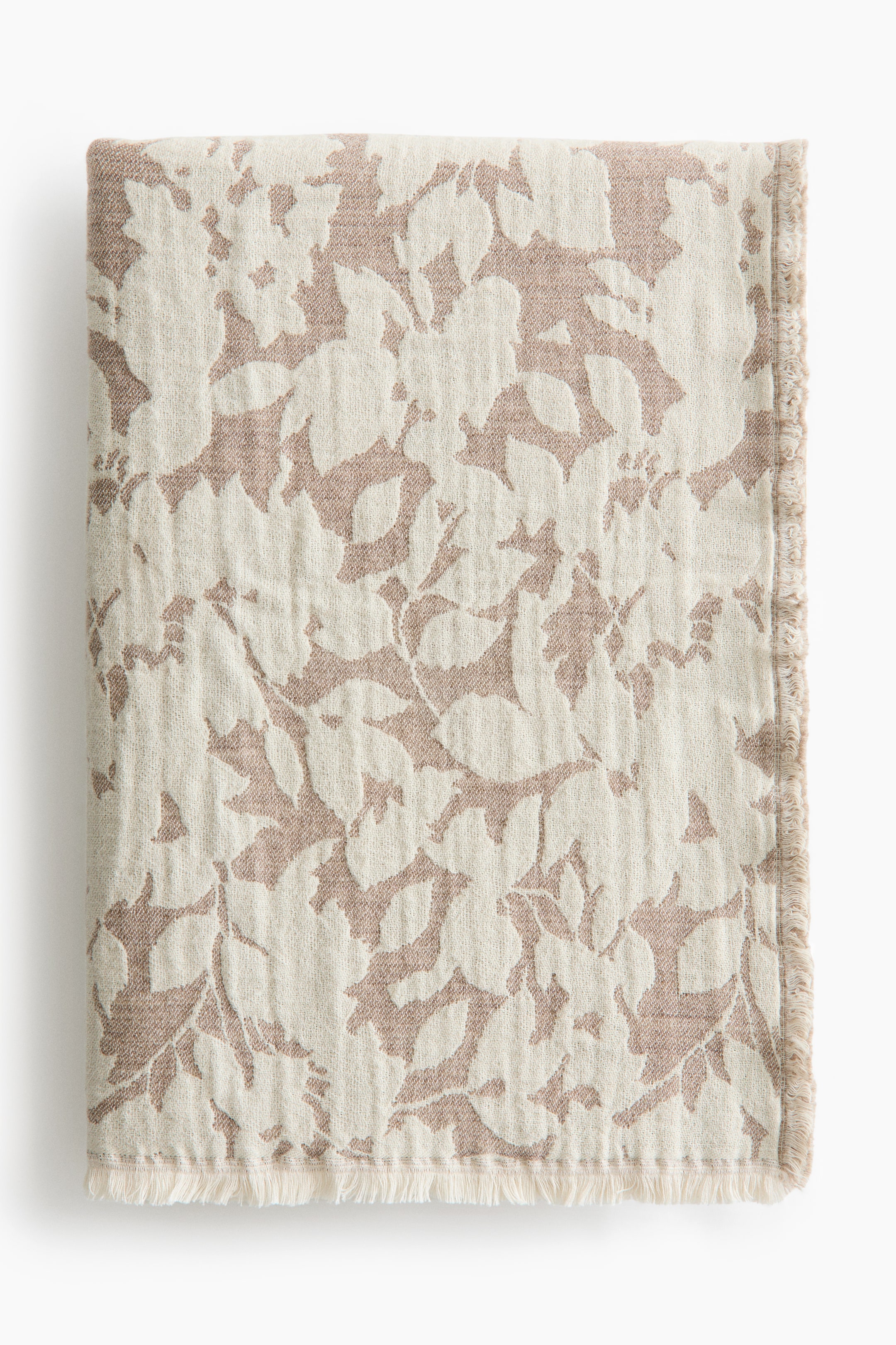 View larger image: A neatly folded blanket showcases an intricate floral texture in light brown, set against a cream backdrop. It boasts fringed edges on its right and bottom sides.