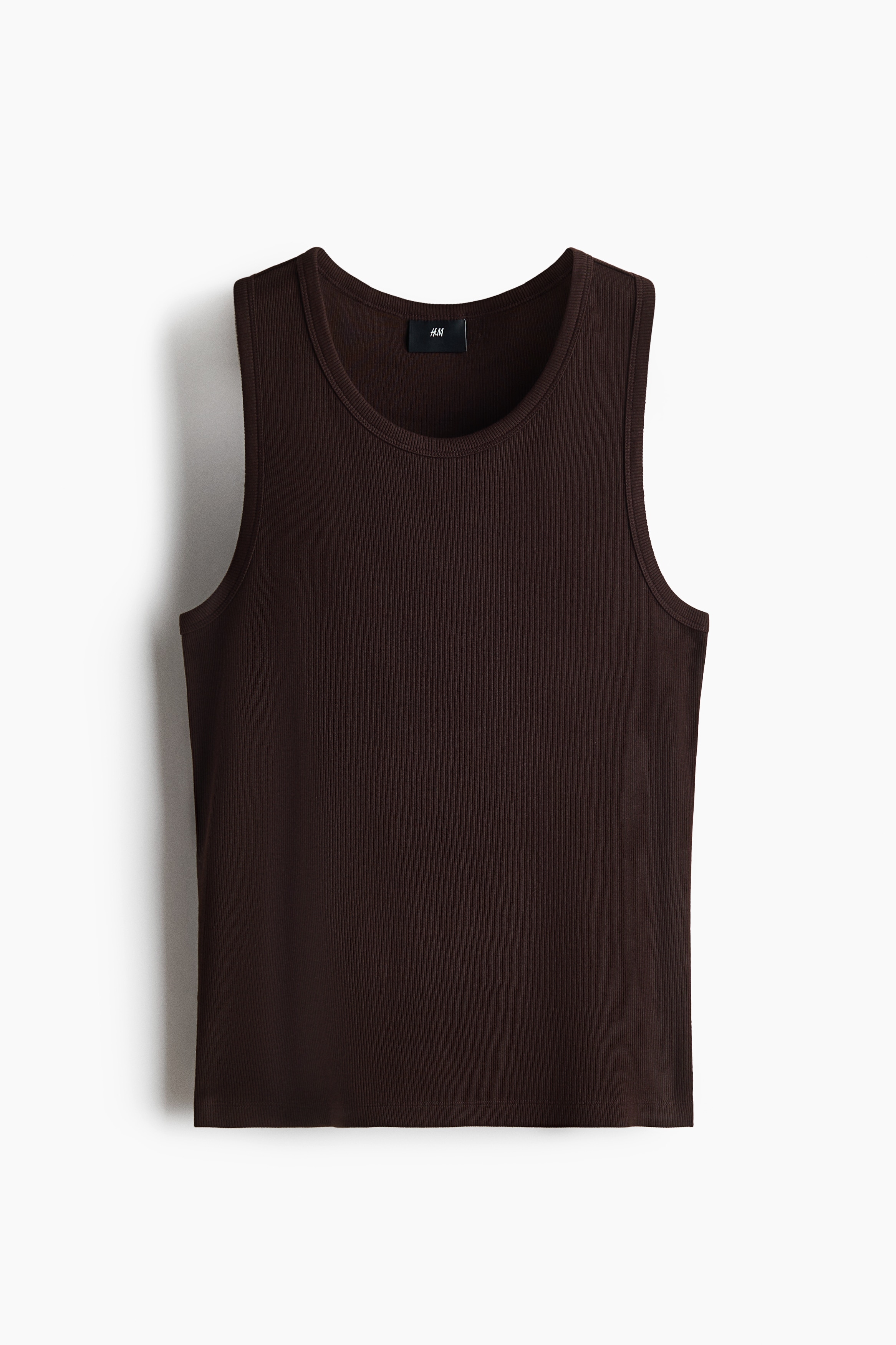 A dark brown, ribbed cotton tank top with a round neckline is being worn by the model. The slim-fit tank is tucked into black trousers secured with a braided belt.