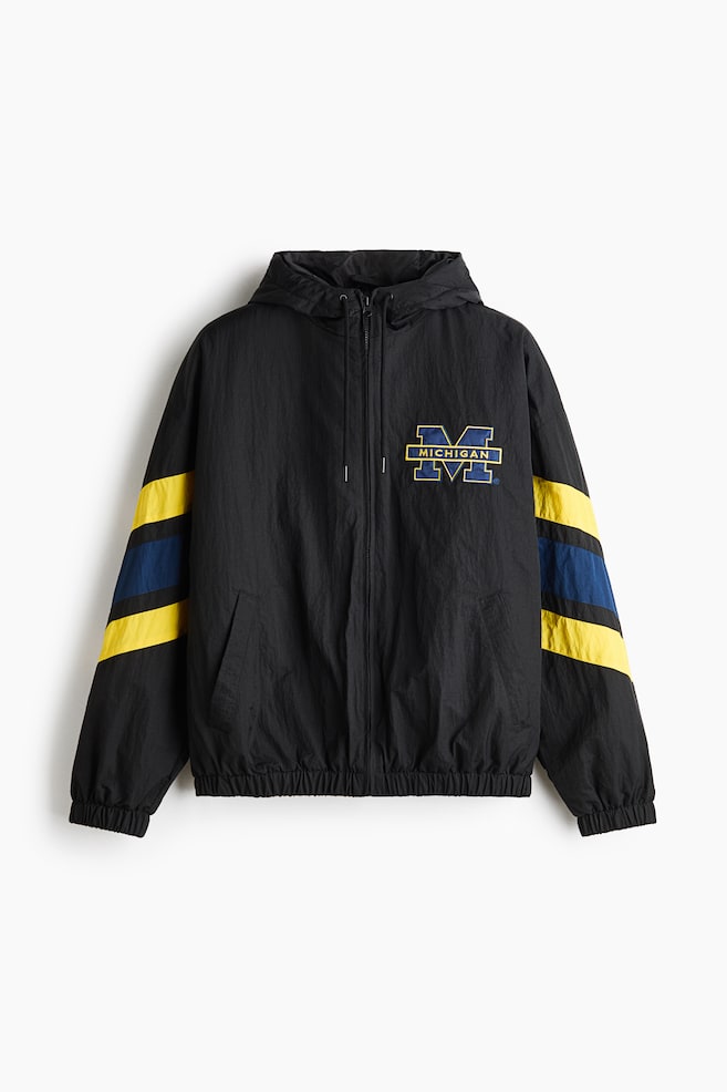 Dit zwarte windjack met capuchon is van voren afgebeeld en heeft een capuchon met trekkoord, een ritssluiting over de volledige lengte, schuine paspelzakken en een elastische zoom en manchetten. Op de linkerborst staat een blauw en geel âMICHIGANâ-logo, met brede diagonale gele en blauwe strepen die om de mouwen heen lopen. - 1