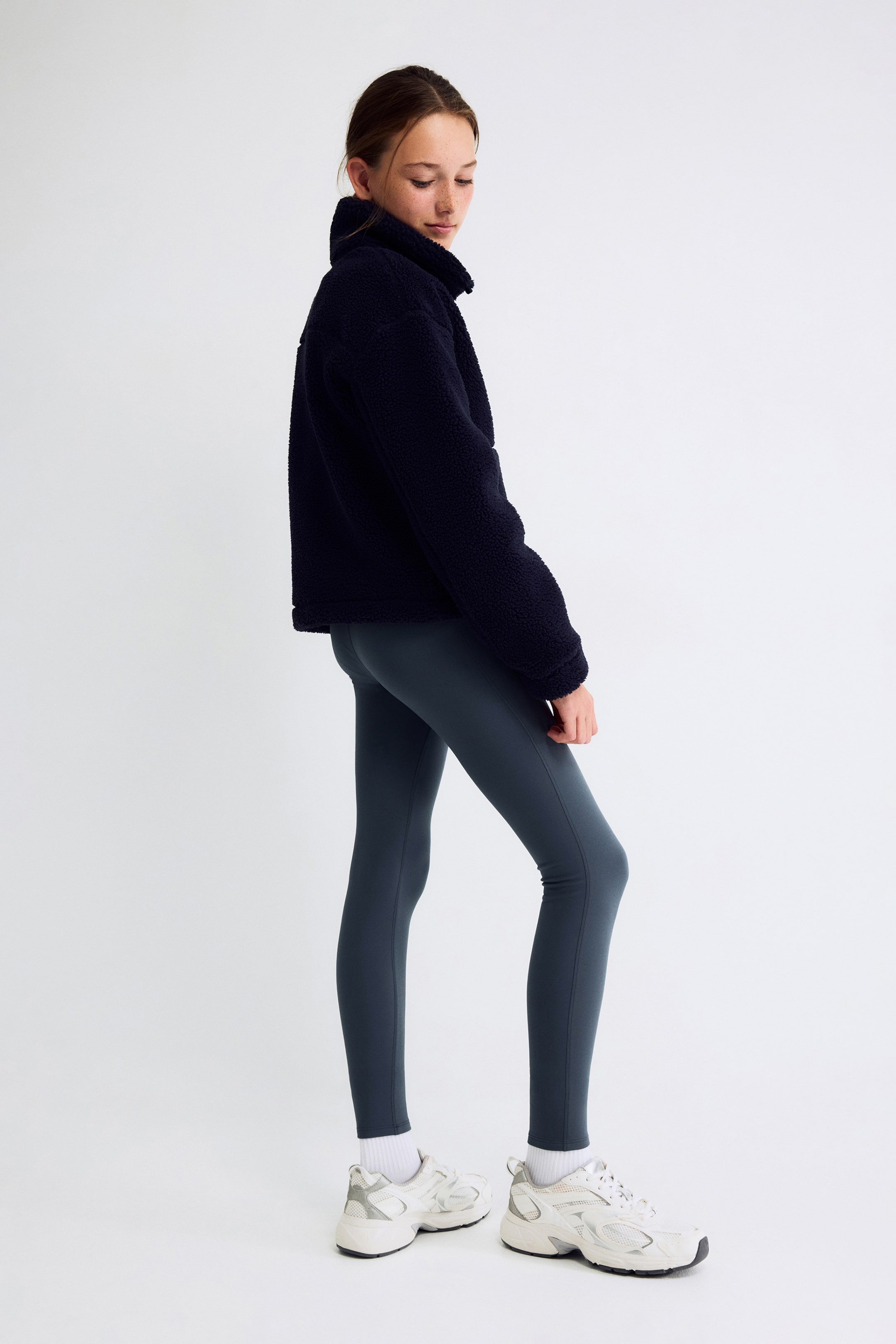 Grotere afbeelding bekijken: Een marineblauwe sherpa-fleecejas, naar links gericht, met een hoge opstaande kraag en een relaxte, licht cropped pasvorm. Ze wordt gecombineerd met nauwsluitende, donkergrijze enkellange leggings die een strakke, atletische look benadrukken.