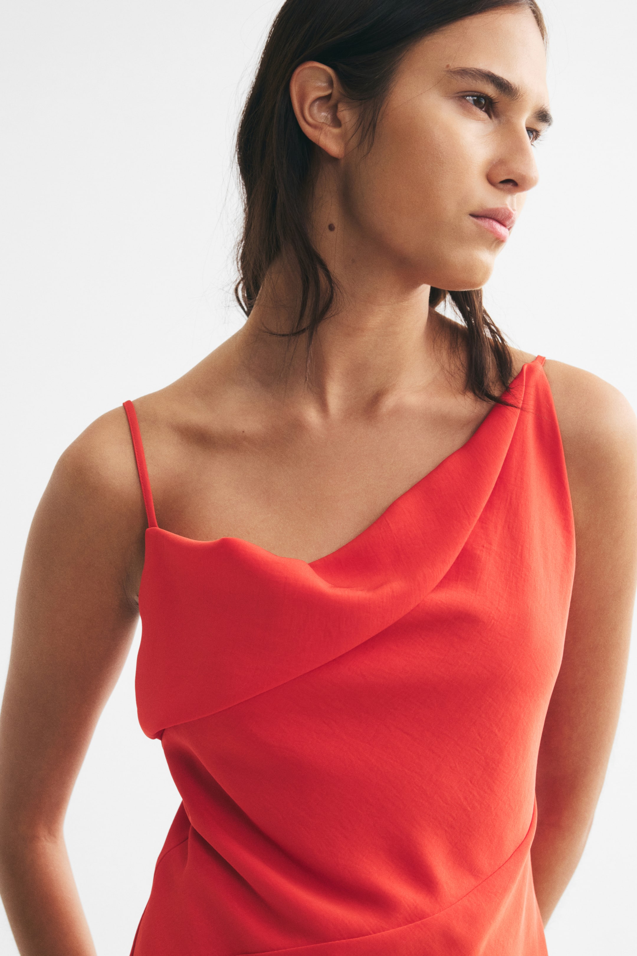 View larger image: The model is wearing a red woven top with an asymmetric draped neckline and a thin shoulder strap. The top features an asymmetric foldover detail at the front and a diagonal seam visible at the waist.