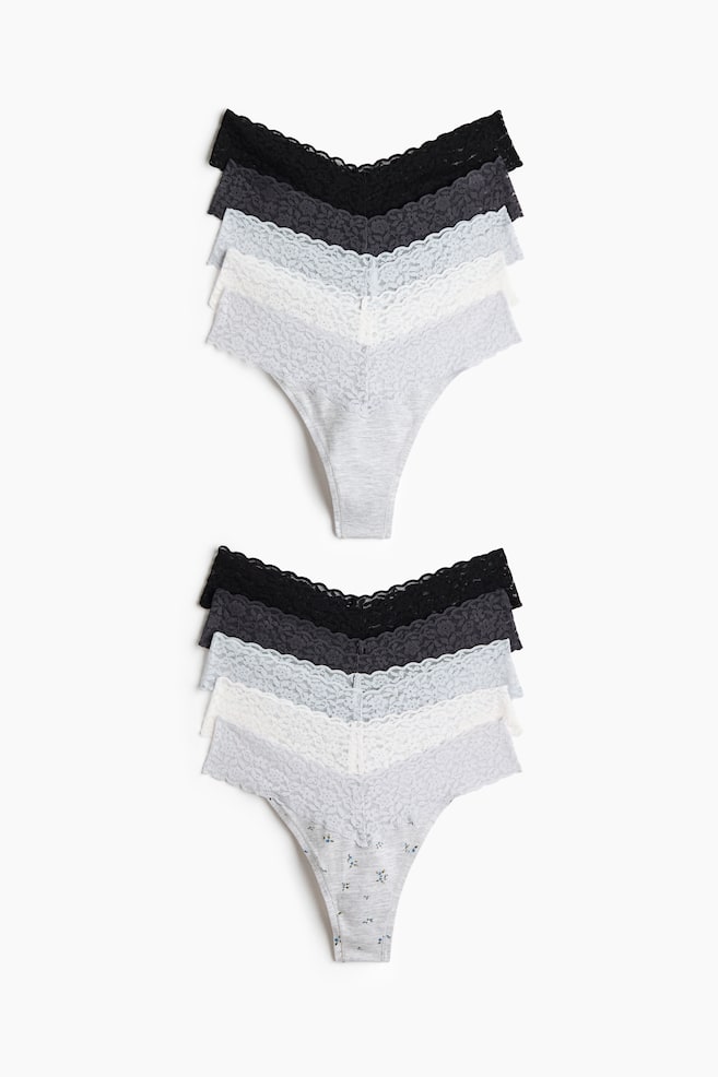 Two stacks of low-rise lace G-strings are shown from the front in a variety of colours, including black, dark grey, light blue, white and heather grey. Each pair features a V-shaped lace waistband and a lined fabric gusset, with the bottom pair displaying a small floral print. - 1