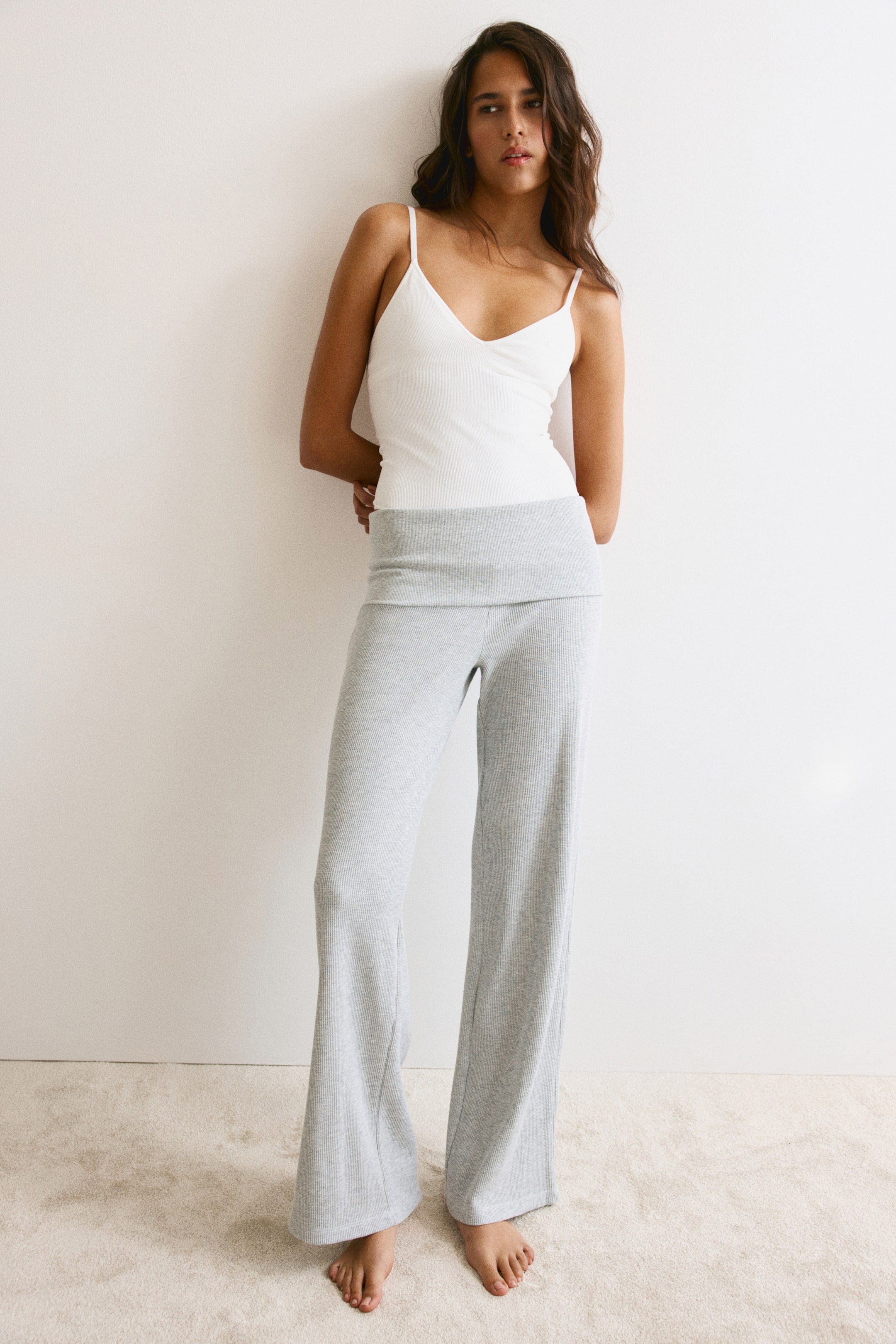 Agrandir l'image: Le mannequin porte un pantalon de pyjama en jersey gaufré gris clair, doté d’une large ceinture rabattable et de jambes droites. Le pantalon est associé à un caraco blanc à fines bretelles, glissé sous la ceinture rabattable.