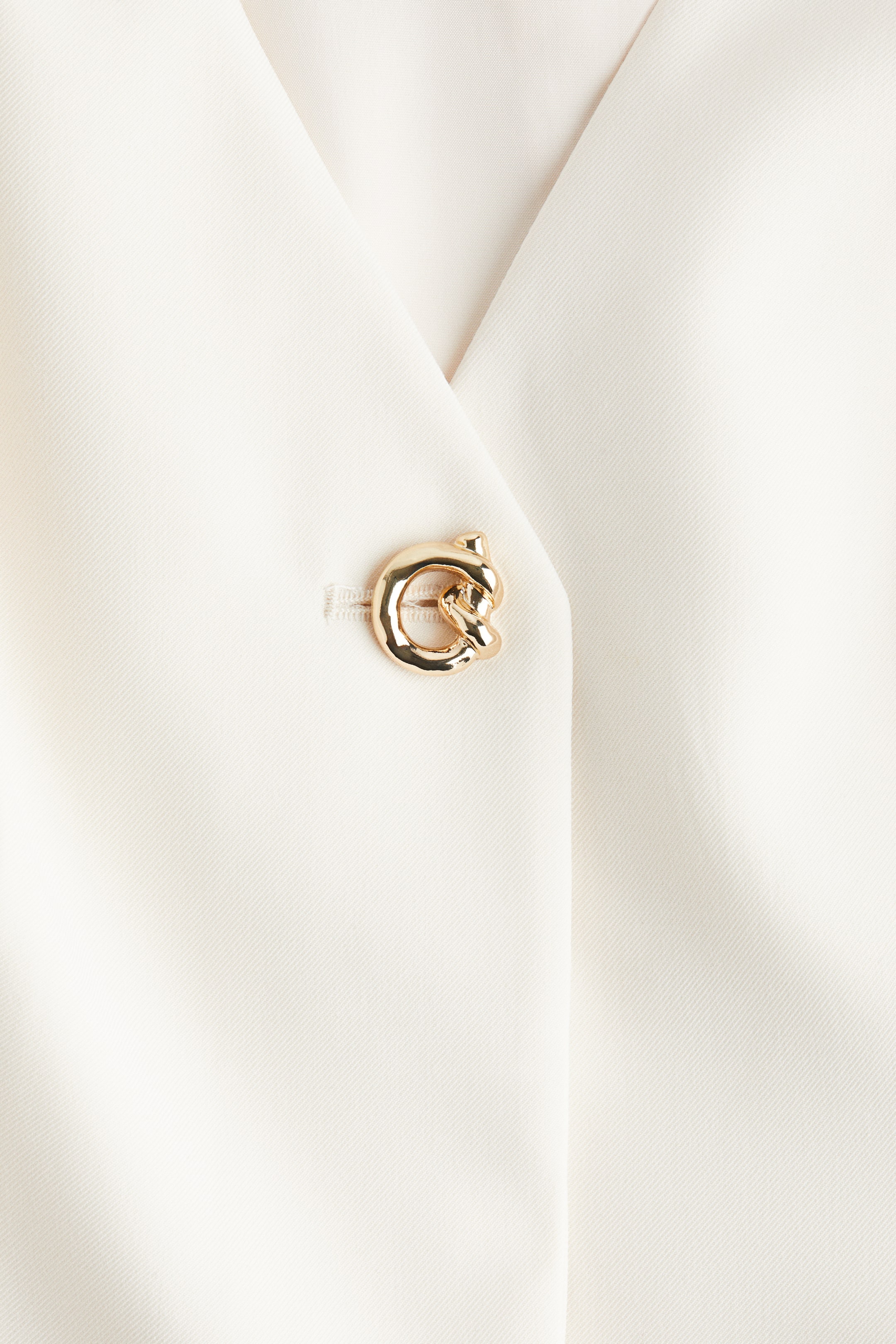 Ampliar la imagen: Una prenda de color crema presenta un cierre frontal cruzado con un escote en V profundo. Un botón prominente en forma de nudo de tono dorado está sujetando la prenda a través de un ojal horizontal.