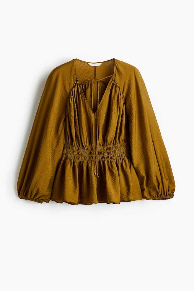 Parte delantera de una blusa oversize de color verde oliva en un tejido texturizado, con un escote en forma de V profundo y lazos estrechos en la parte superior, costuras de los hombros fruncidas y mangas largas raglán abullonadas con puños elásticos estrechos, y nido de abeja en la cintura que crea un dobladillo peplum fruncido. - 1