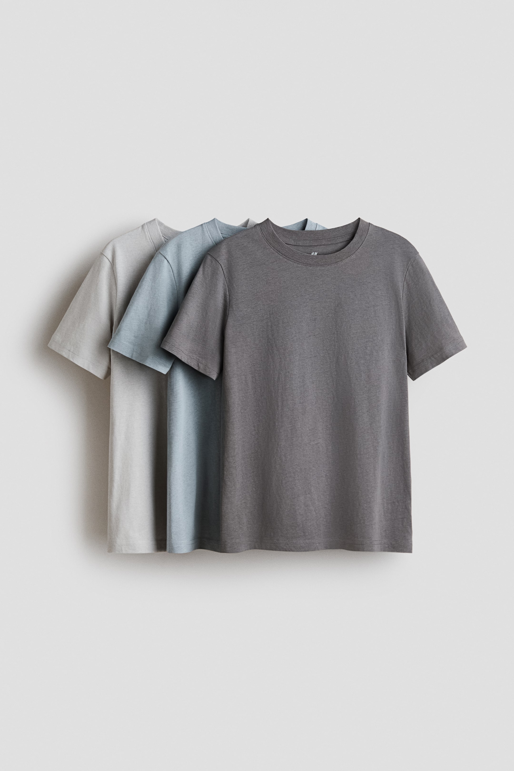 Xem ảnh lớn hơn: Áo thun cổ tròn bằng chất liệu cotton jersey mềm mại được trưng bày với góc nhìn từ phía trước, sắp xếp theo kiểu xếp lớp. Các màu sắc hiển thị là xám nhạt, xanh nhạt và xám than, mỗi chiếc đều có tay ngắn và cổ áo bo gân.