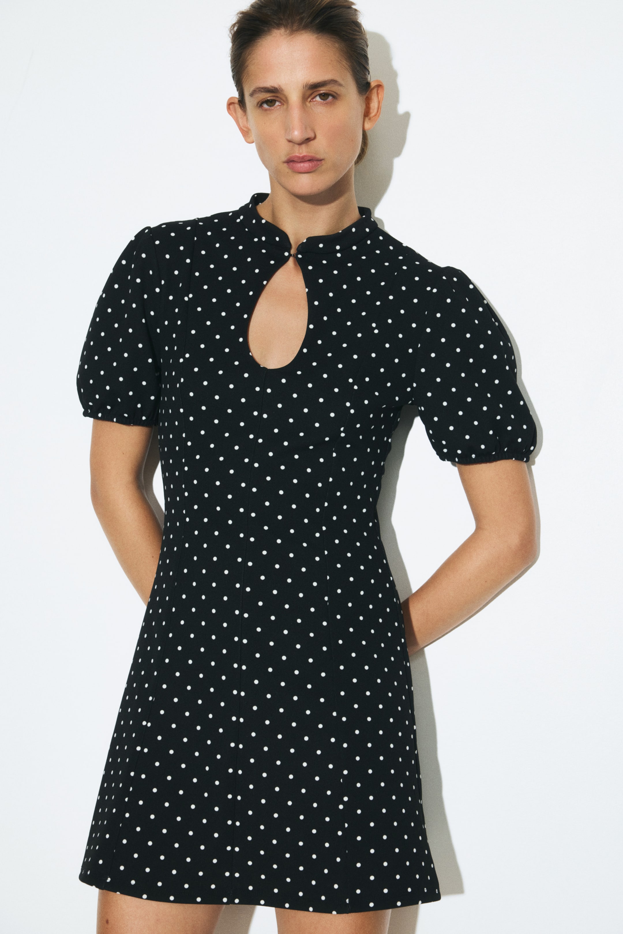 Ampliar la imagen: La modelo lleva puesto un vestido corto negro con lunares blancos. El vestido presenta un escote redondo con abertura tipo ojo de cerradura en el frente, mangas cortas abullonadas y un corpiño ajustado con costuras verticales visibles.