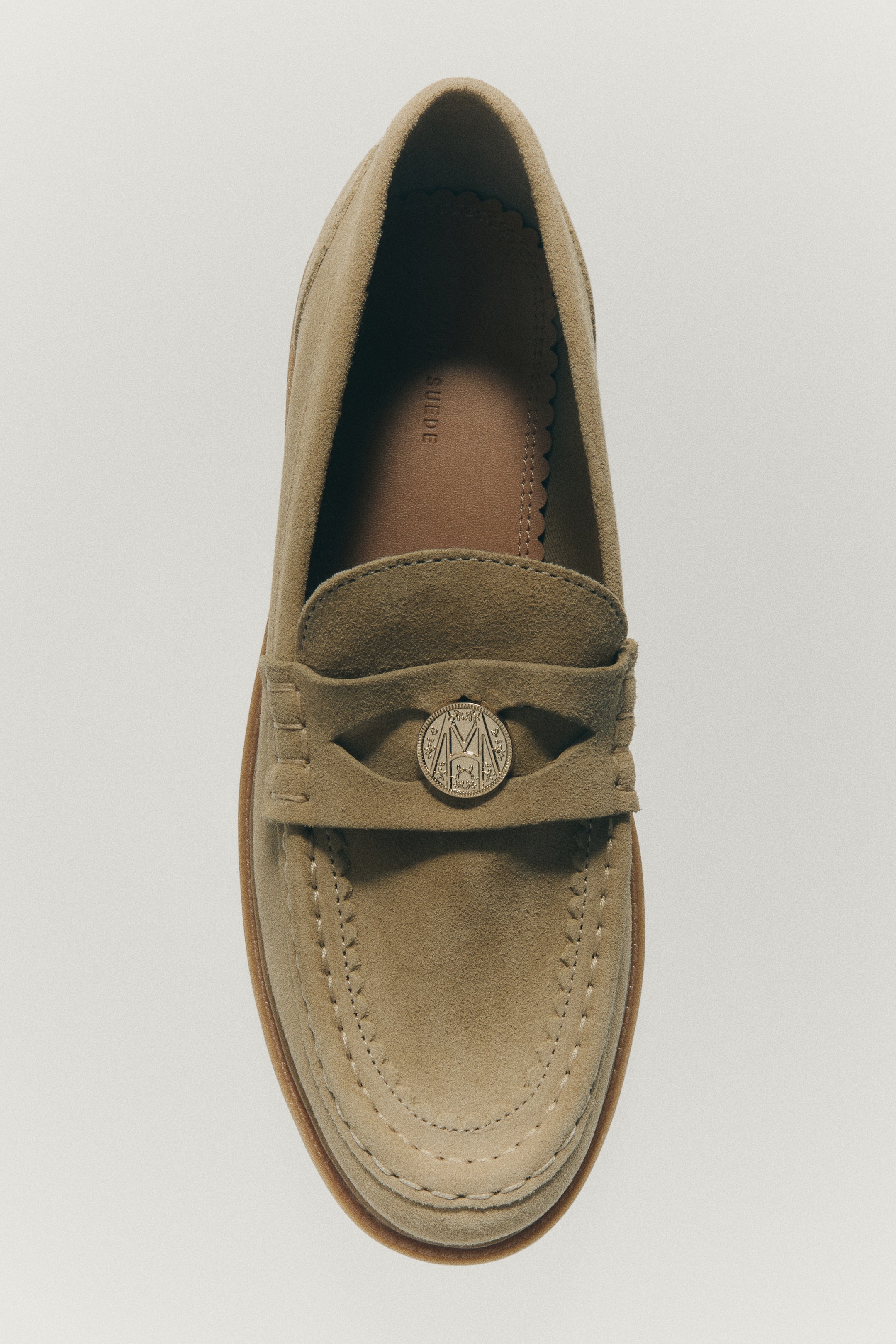View larger image: A beige split-suede loafer is shown in a top-down view, featuring a stitched moccasin seam and a penny keeper strap across the front. The strap displays a decorative gold-toned metal coin detail at the centre, while the inside shows a tan lining.