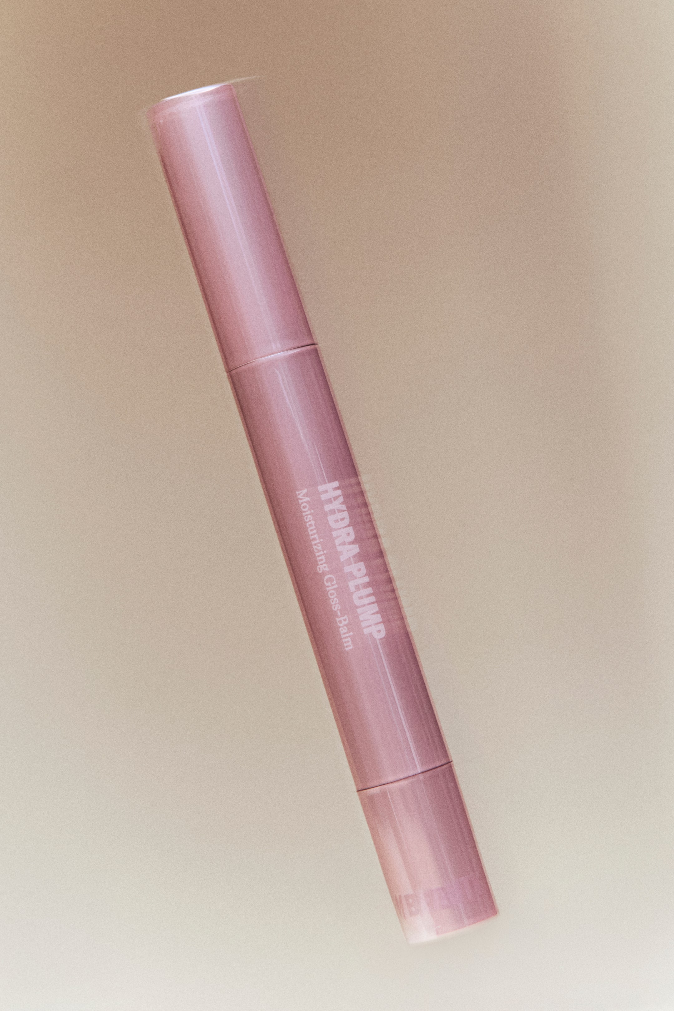 View larger image: A cylindrical, muted pink lip gloss balm is angled diagonally upwards to the right. The words 'HYDRA PLUMP Moisturizing Gloss-Balm' appear vertically along its smooth, slightly glossy surface.
