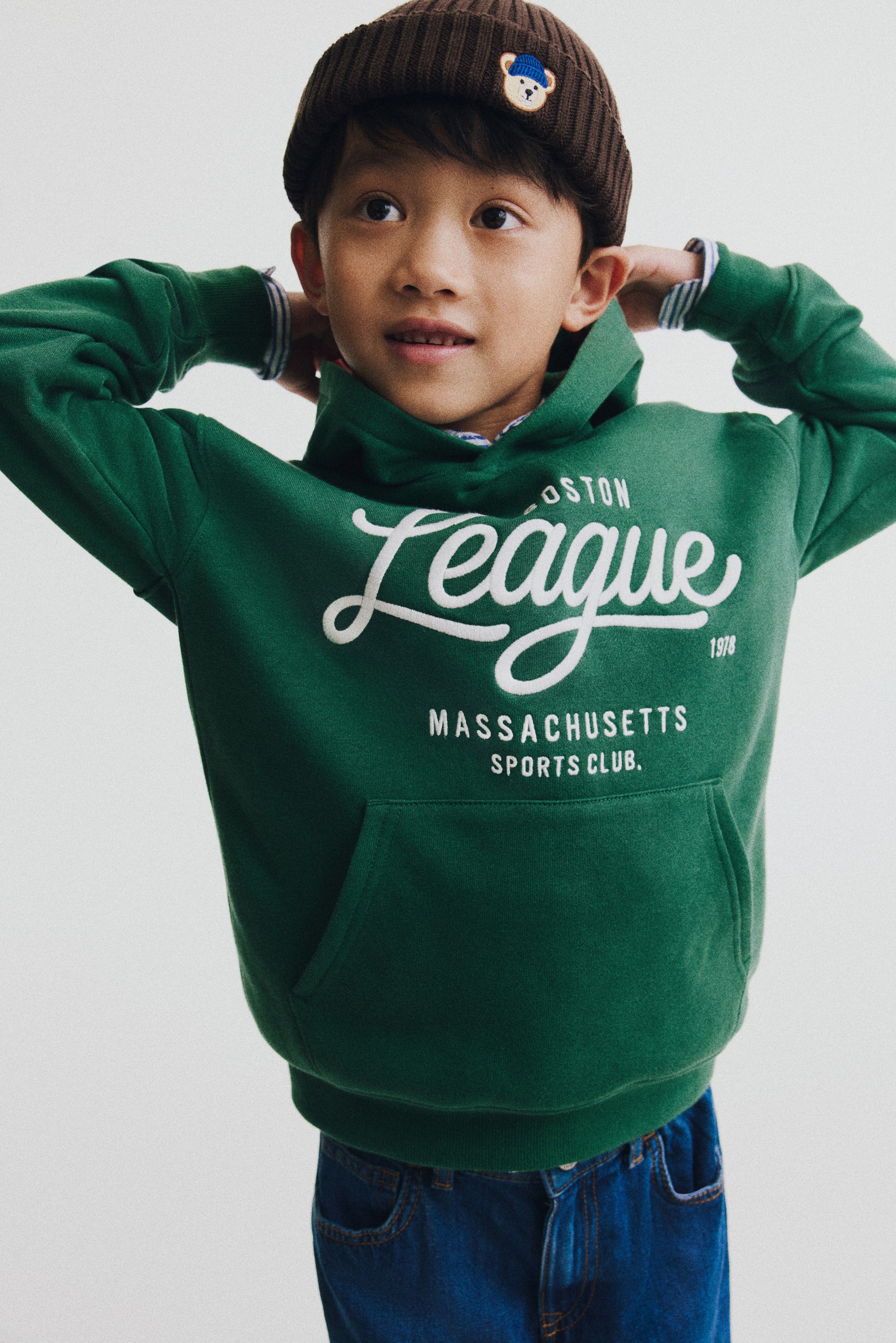 View larger image: A green loose-fit hoodie is shown from the front, featuring a wrap-front hood, dropped shoulders, and a white graphic print on the chest. The hoodie showcases a kangaroo pocket and a wide ribbed hem, with striped cuffs from an underlayer peeking out at the wrists.