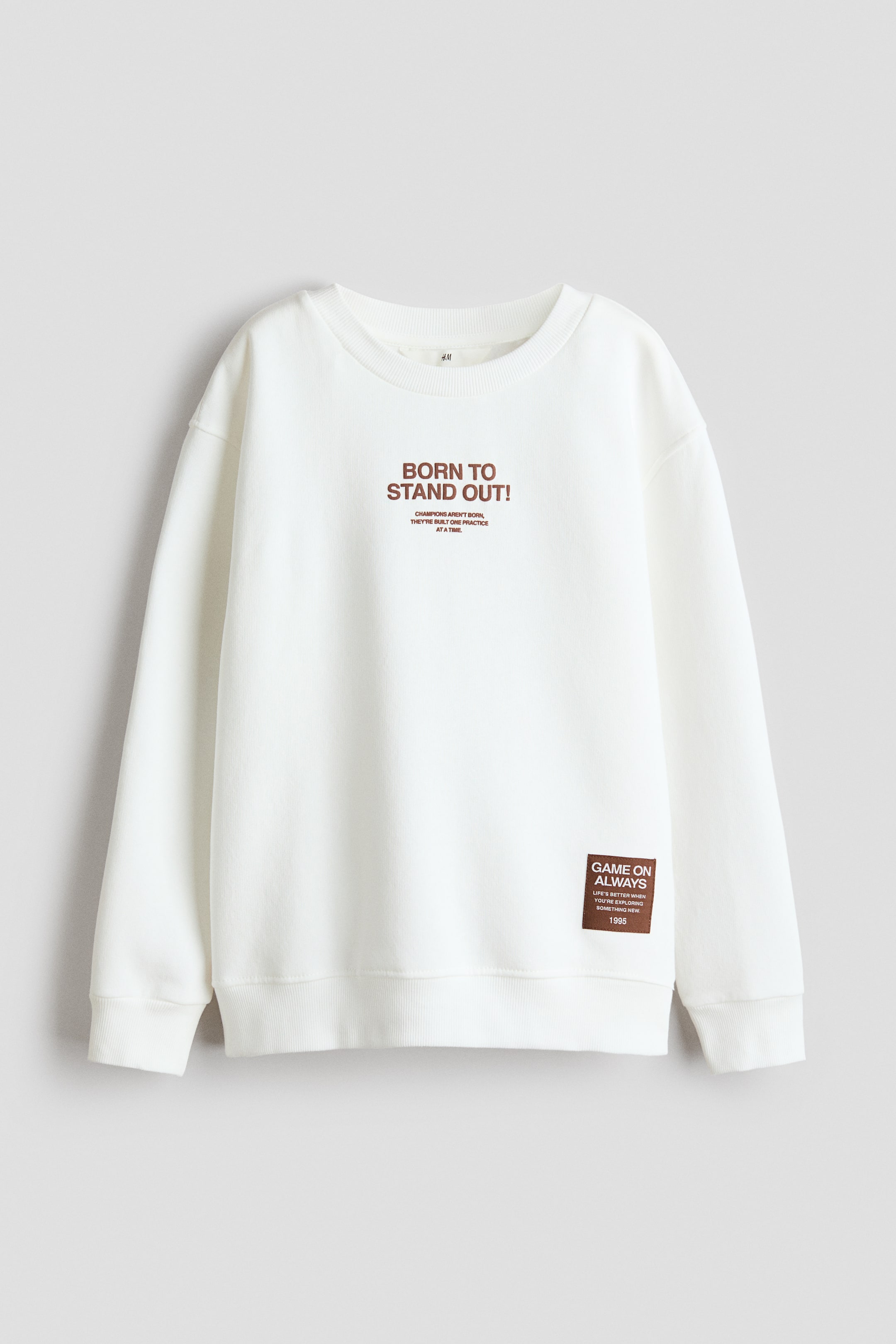 Ver imagen más grande: Una sudadera blanca de corte holgado está de frente, con un cuello redondo ribeteado, hombros caídos, mangas largas con puños acanalados y un dobladillo acanalado. Muestra un motivo de impresión marrón en el pecho que dice "¡NACIDO PARA DESTACAR!" y una impresión rectangular marrón en la parte inferior derecha.