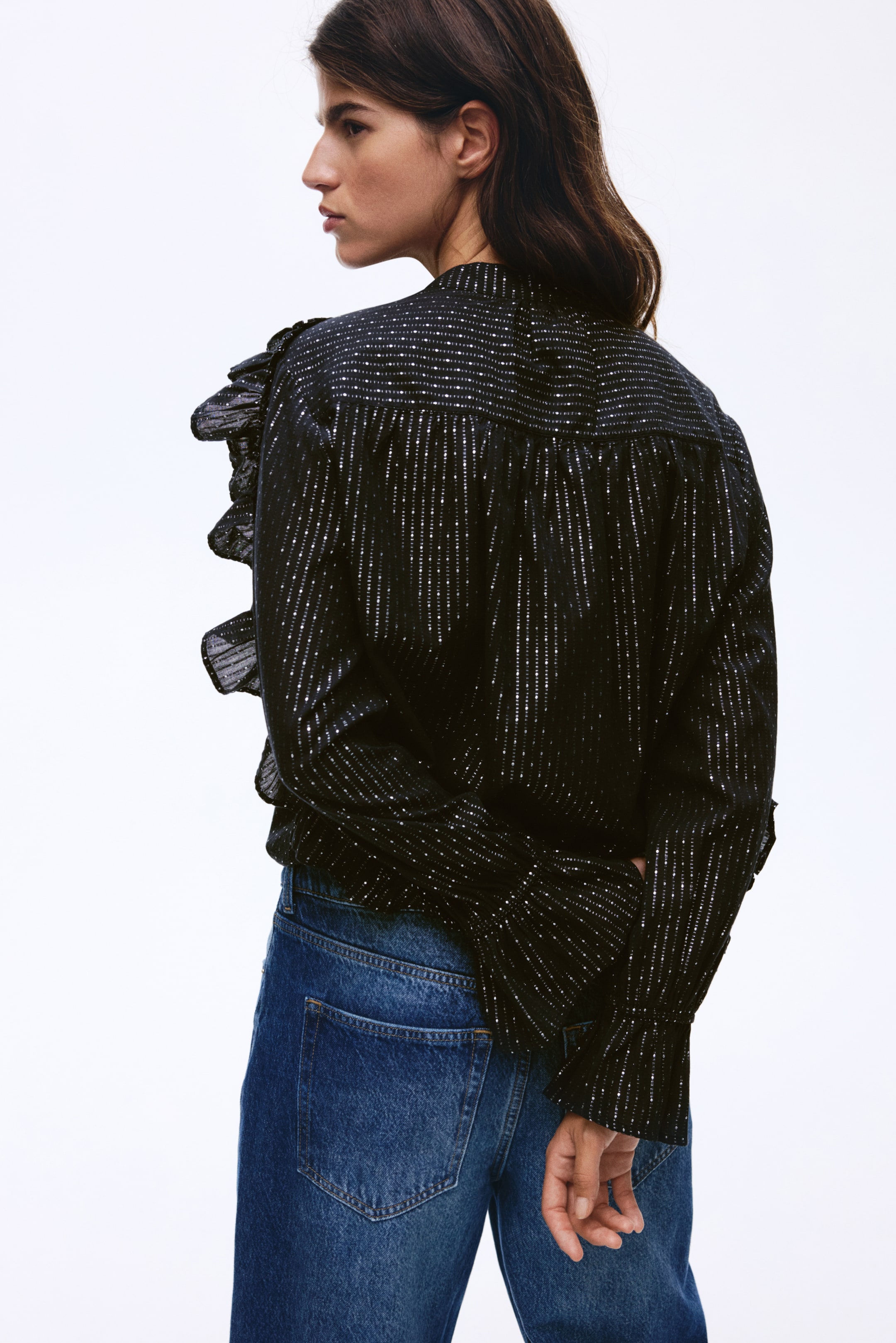 Agrandir l'image: Une blouse sombre, ornée de rayures verticales scintillantes, est portée et vue du côté arrière droit, mettant en avant un empiècement smocké sur le haut du dos. Des volants décorent l'épaule droite, et les manches longues se terminent par des poignets smockés.