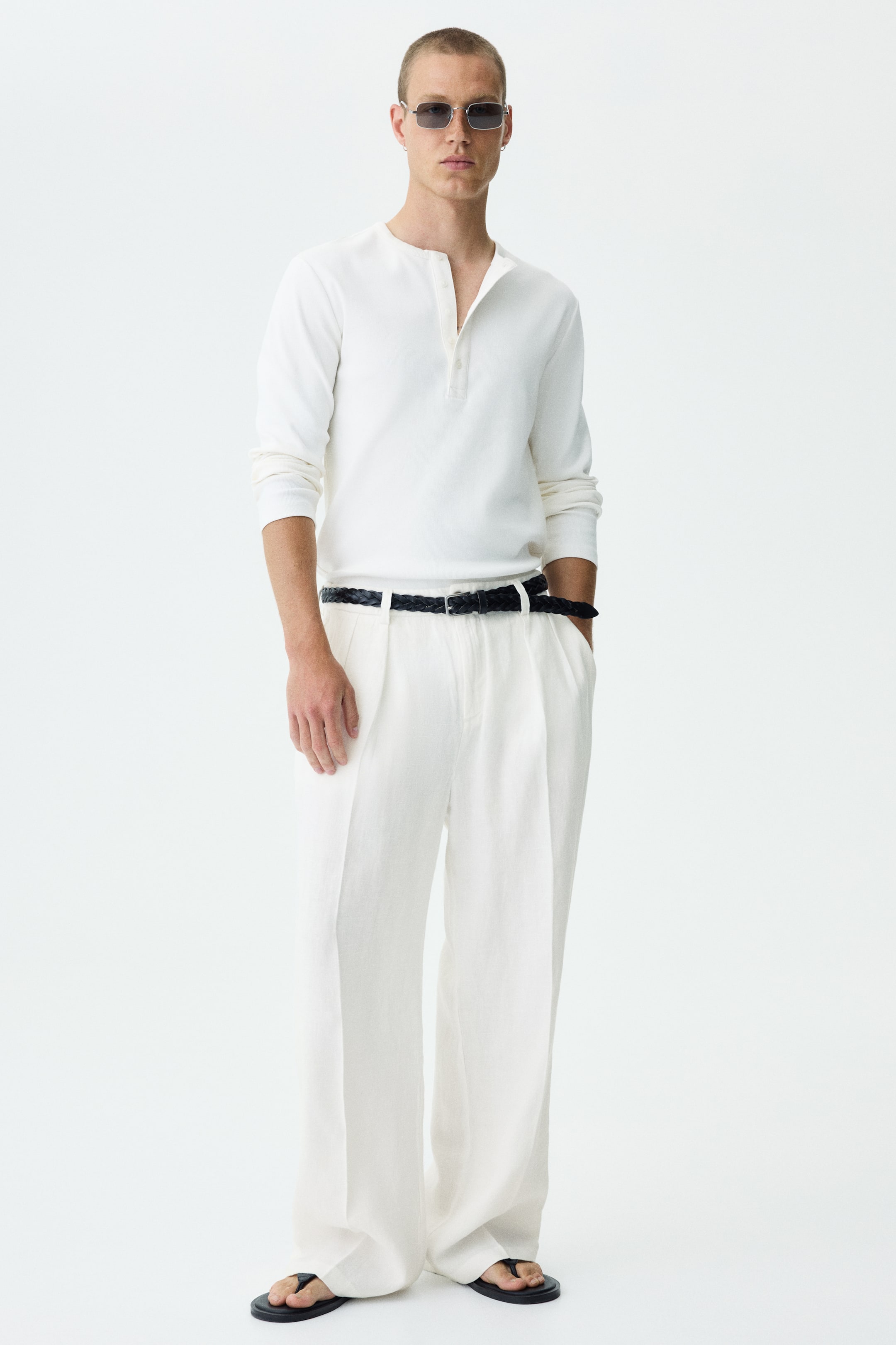 View larger image: The ensemble showcases a white long-sleeve henley shirt with an open button placket, paired with white wide-leg pleated pants. A black braided belt cinches the waist of the pants, while black flip-flops complete the look from the front.