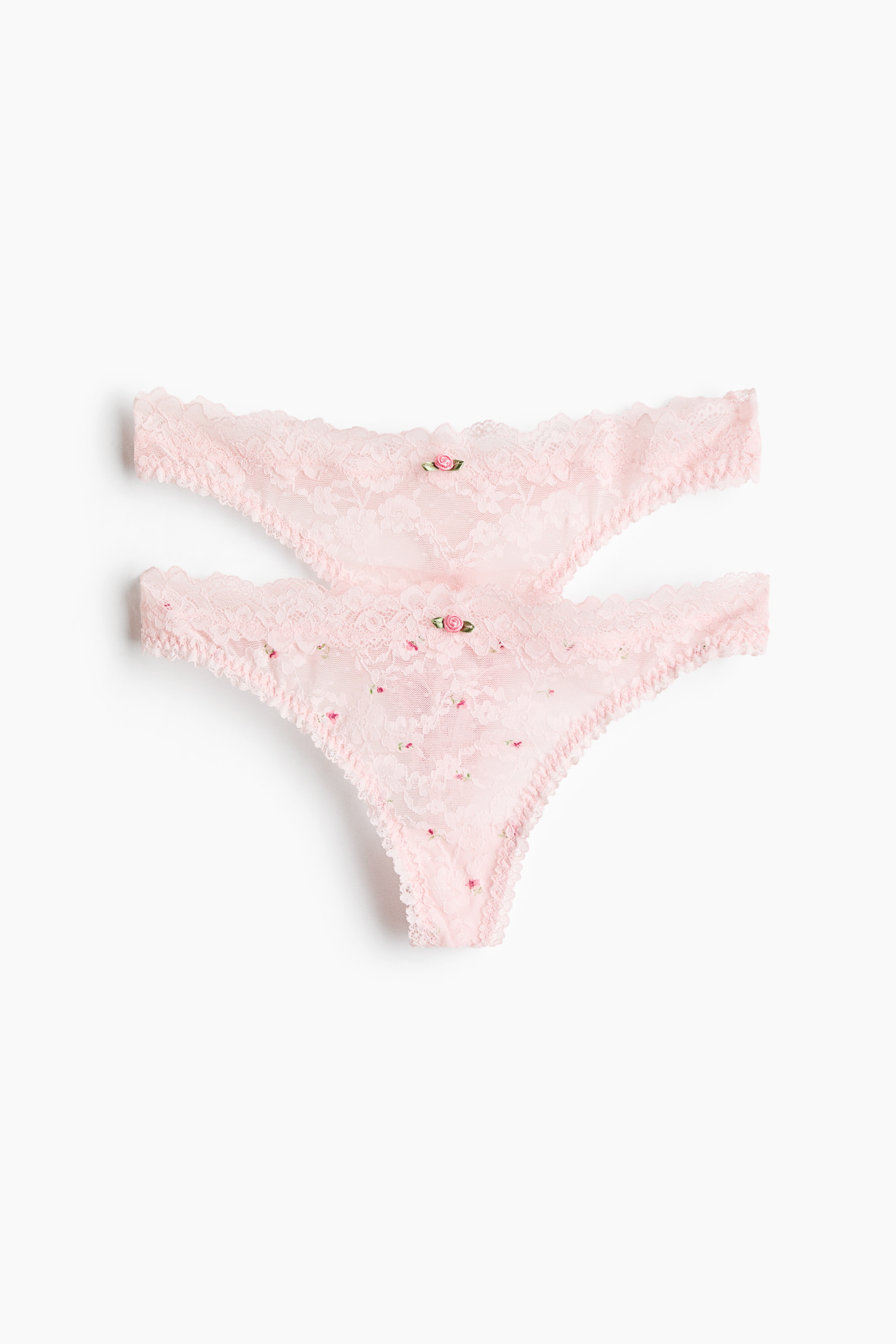 Lace G-strings featuring a low-rise waist, scalloped edges, and a pink floral pattern are being worn by the model. The G-strings are paired with a matching pink lace underwire bra with similar floral details.