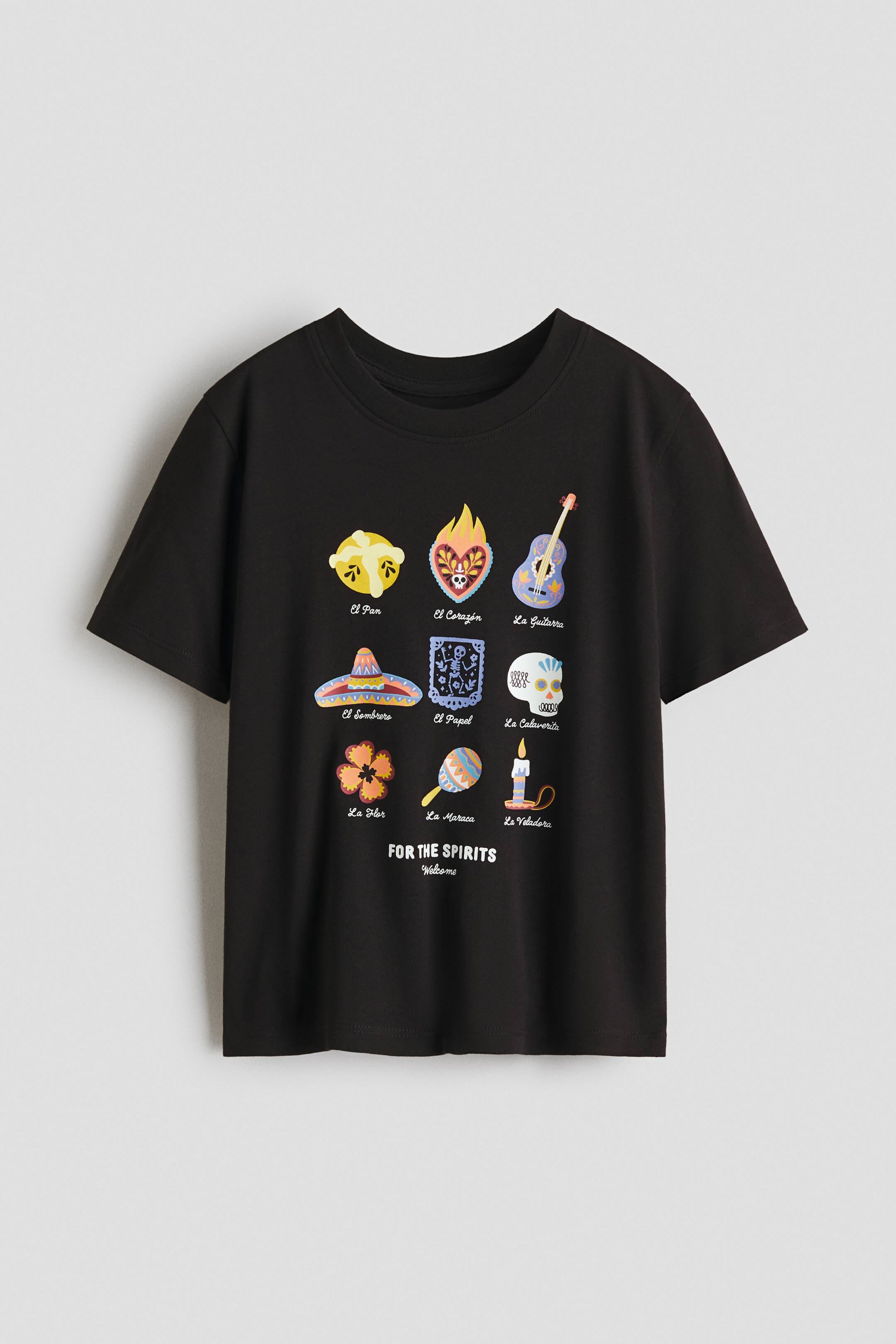 View larger image: A black short-sleeve t-shirt is shown from the front, featuring a crew neck and a vibrant graphic print on the front. The design showcases a grid of nine illustrations, including a guitar, a flaming heart, and a sugar skull, each labeled in Spanish, accompanied by the text 'FOR THE SPIRITS Welcome' underneath.