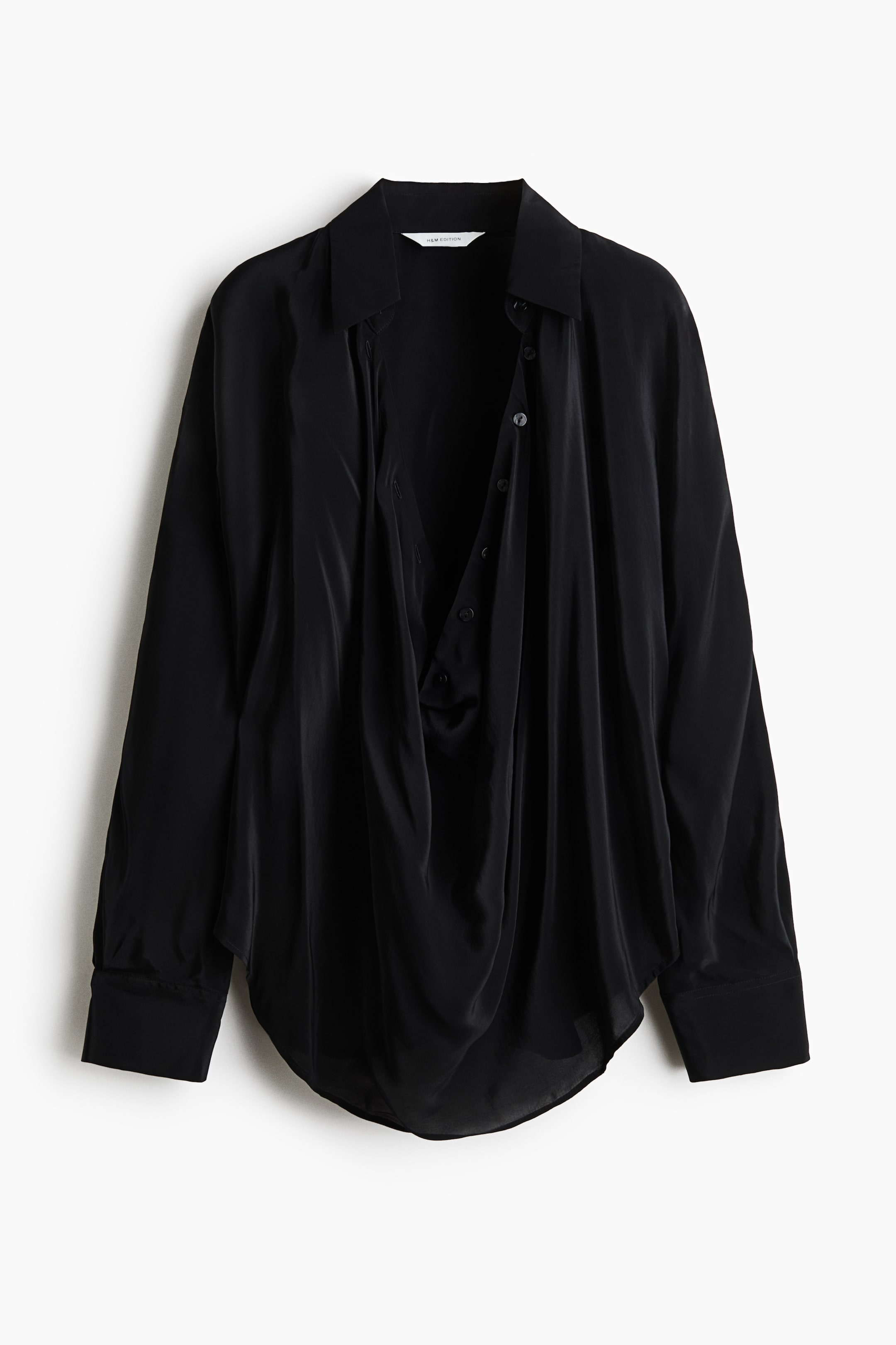View larger image: A front-facing view of an oversized black blouse in draping satin fabric, featuring a collar and a deep, draped front opening with buttons. The blouse features long batwing sleeves with wide button cuffs and a rounded hem.