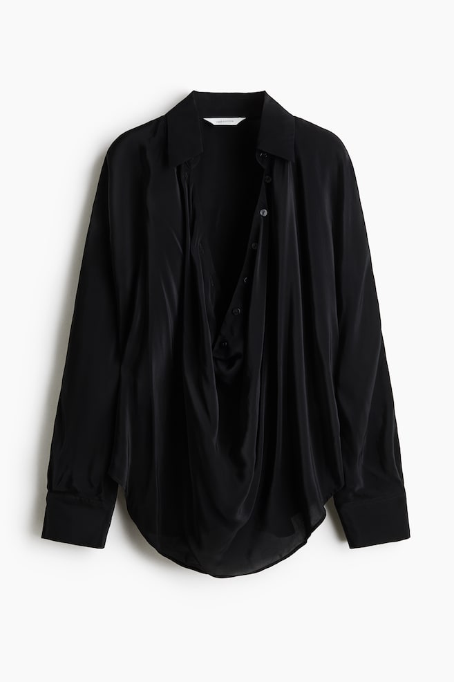 A front-facing view of an oversized black blouse in draping satin fabric, featuring a collar and a deep, draped front opening with buttons. The blouse features long batwing sleeves with wide button cuffs and a rounded hem. - 1