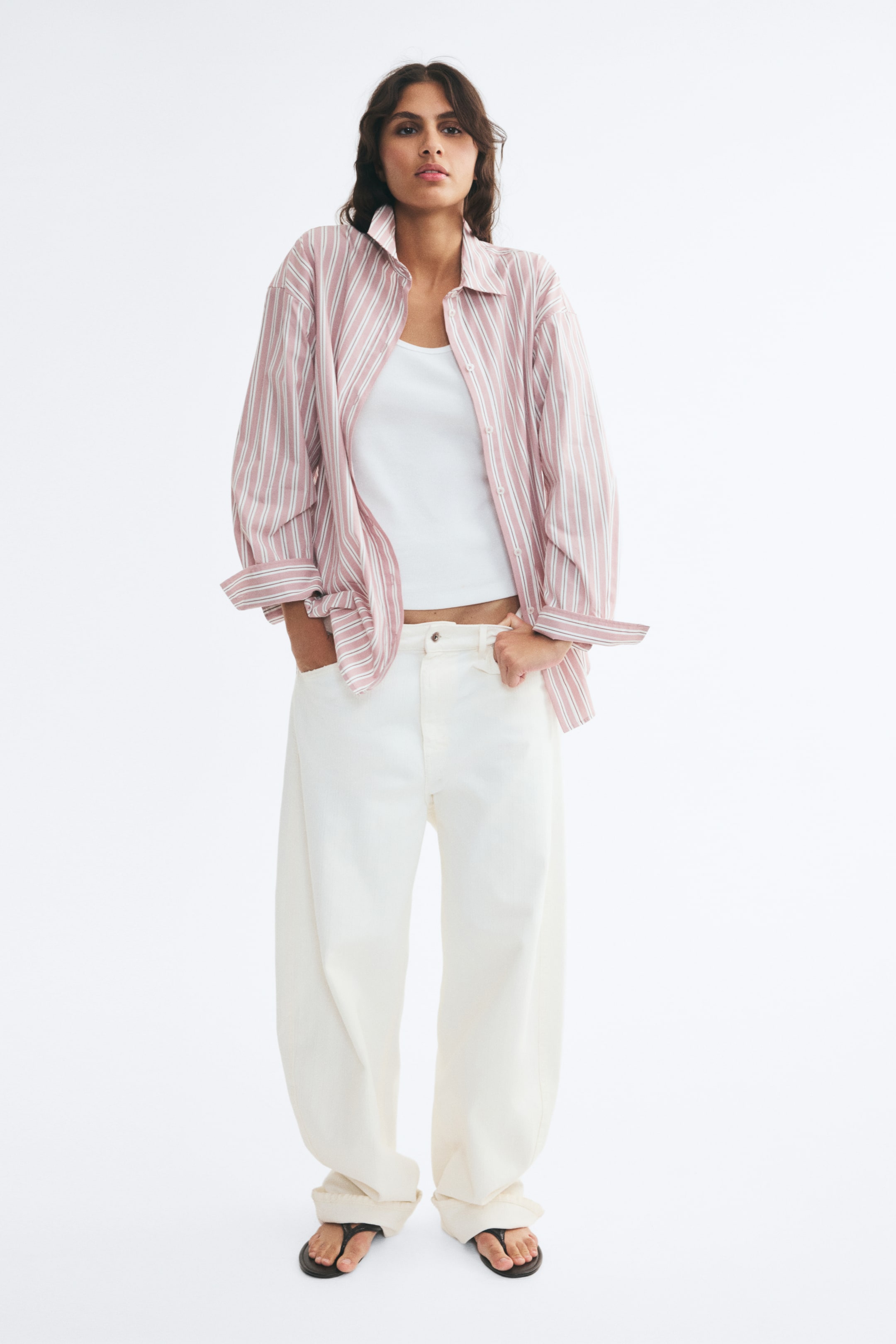 View larger image: An oversized pink-and-white vertically striped cotton shirt is worn open by the model, layered over a white tank top and paired with wide-leg off-white trousers. The shirt features dropped shoulders, a collar, and long sleeves with folded cuffs.