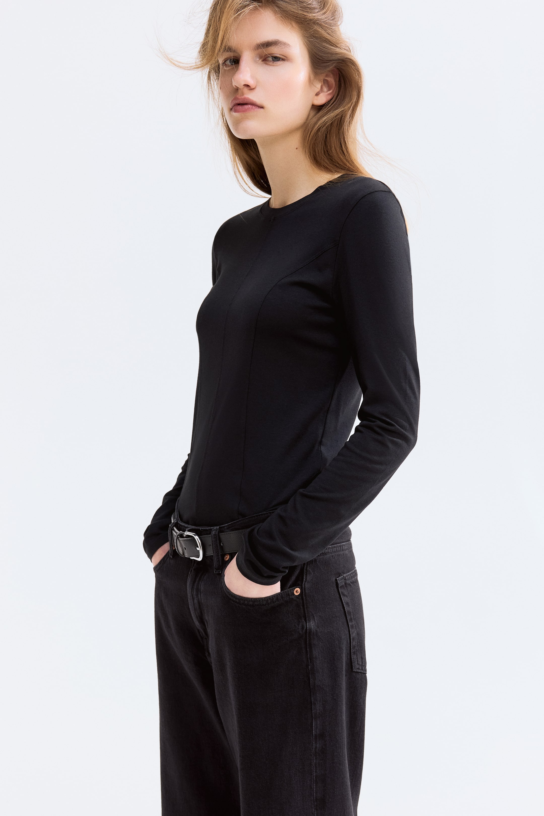 拡大画像を表示: 黒の長袖トップスがわずかに斜めから見える角度で紹介されており、クルーネックとフロントの縦のシェーピングシームが見えるフィット感のある構造を強調しています。ダークジーンズにインしてスタイリングされています。