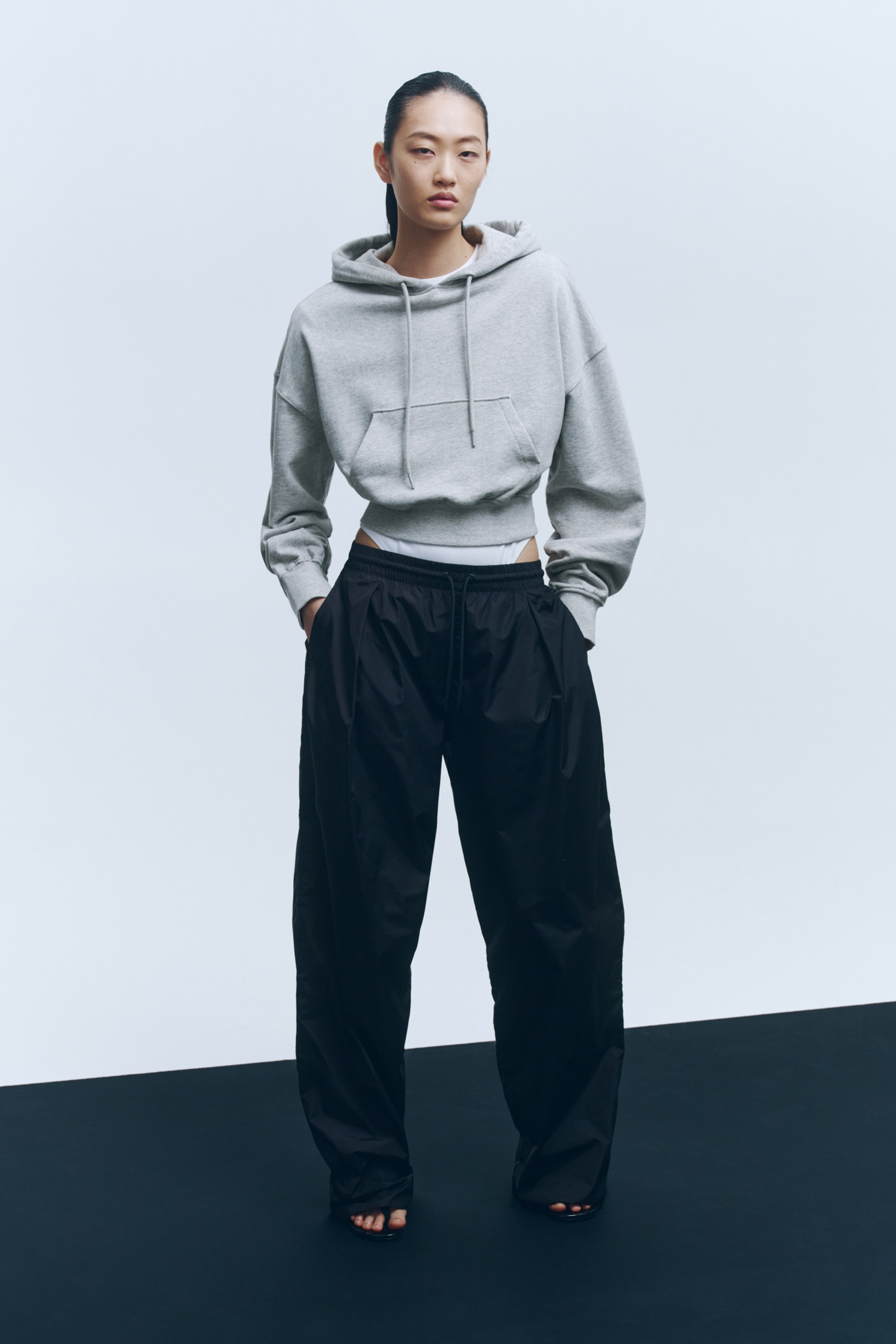 Ver imagem maior: Camisola cropped com capuz em cinzento claro, com cordão ajustável e bolso canguru. A modelo está a usar a camisola, que está sobreposta a uma peça de roupa branca.