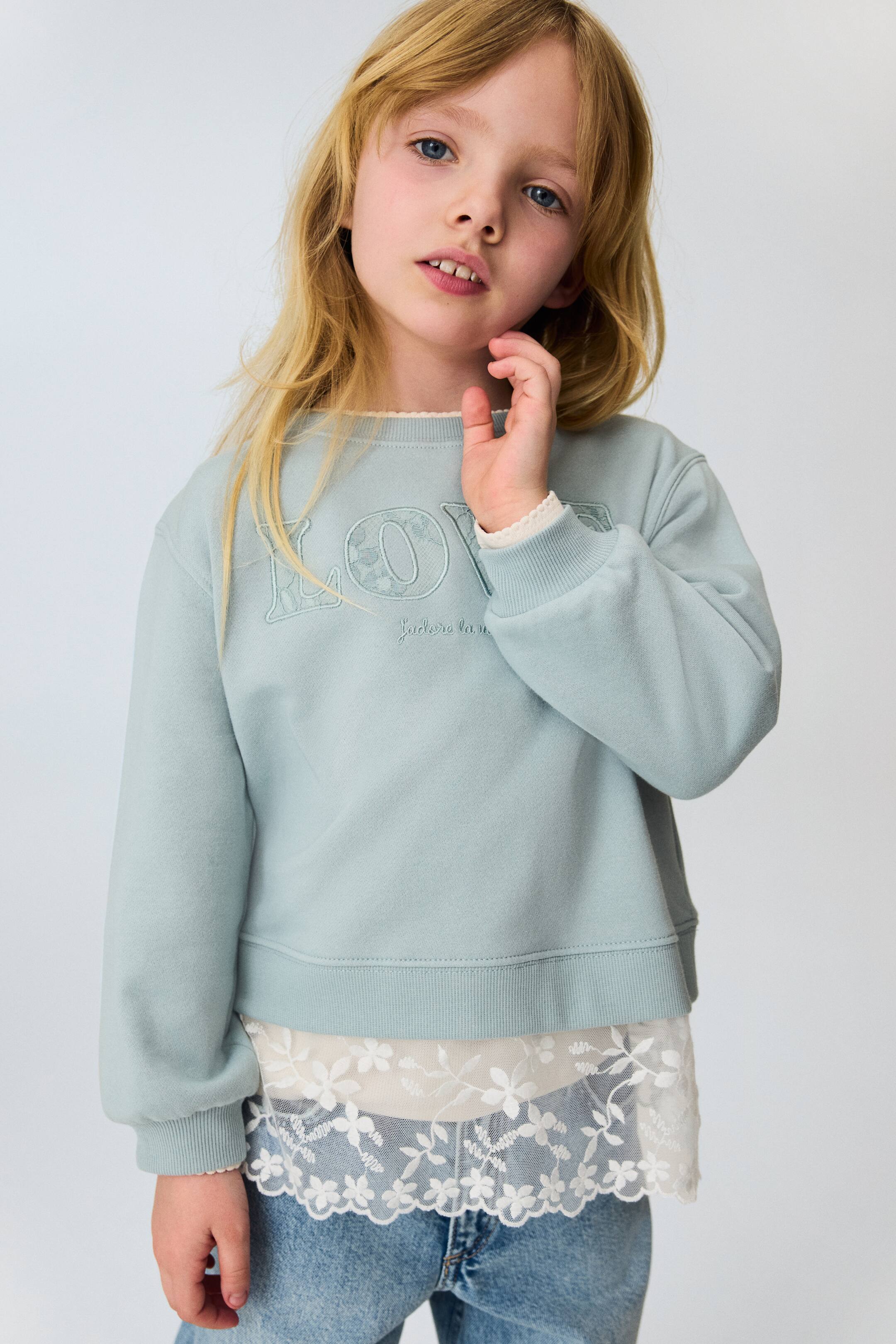 View larger image: A light blue crew-neck sweatshirt features "LOVE" embroidered on the front chest with a lace-patterned fill and "J'adore la vie" text below. It is designed with ribbed cuffs and a ribbed hem, layered over a white top with a ruffled edge visible at the neckline and cuffs, and a white floral lace hem extending below.