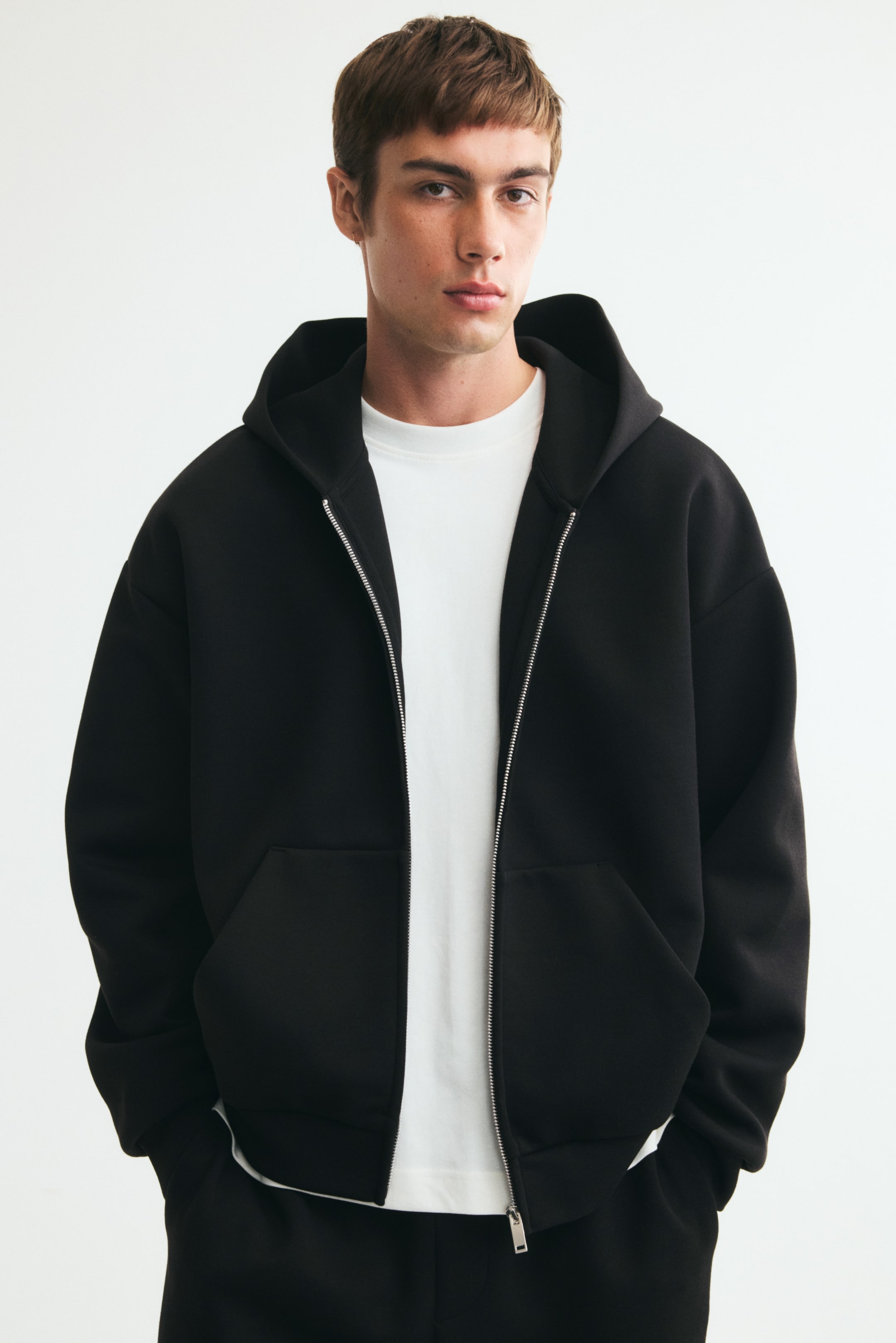 View larger image: A black zip-up hoodie in interlock jersey is styled open on the model over a white crew-neck T-shirt. The hoodie features an oversized silhouette with dropped shoulders, a silver-tone front zipper, front pockets, and a lined hood.