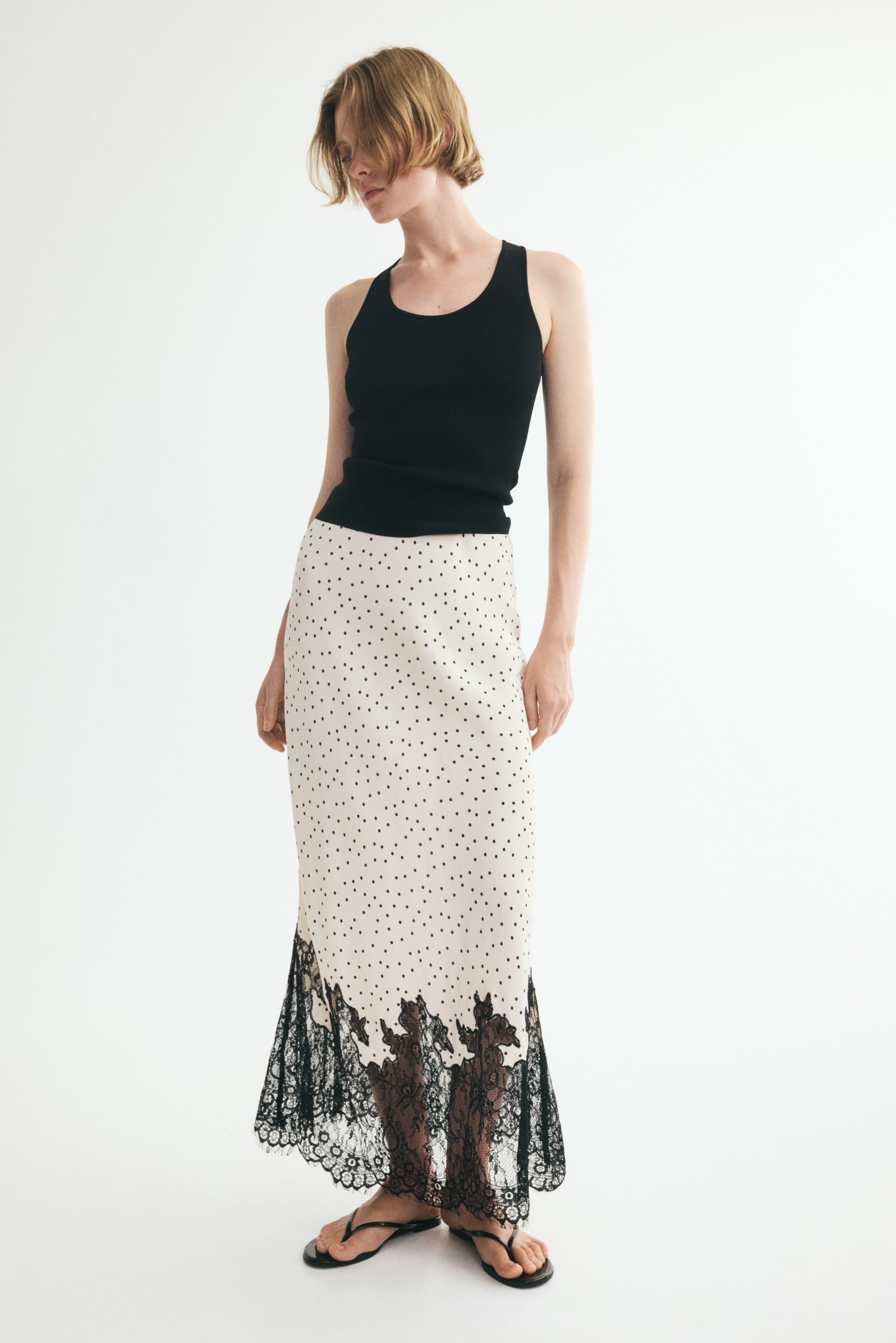 View larger image: A long, fitted, cream-coloured skirt with black polka dots and a wide black lace trim at the hem is being worn by the model. The model is styling the skirt with a black tank top.