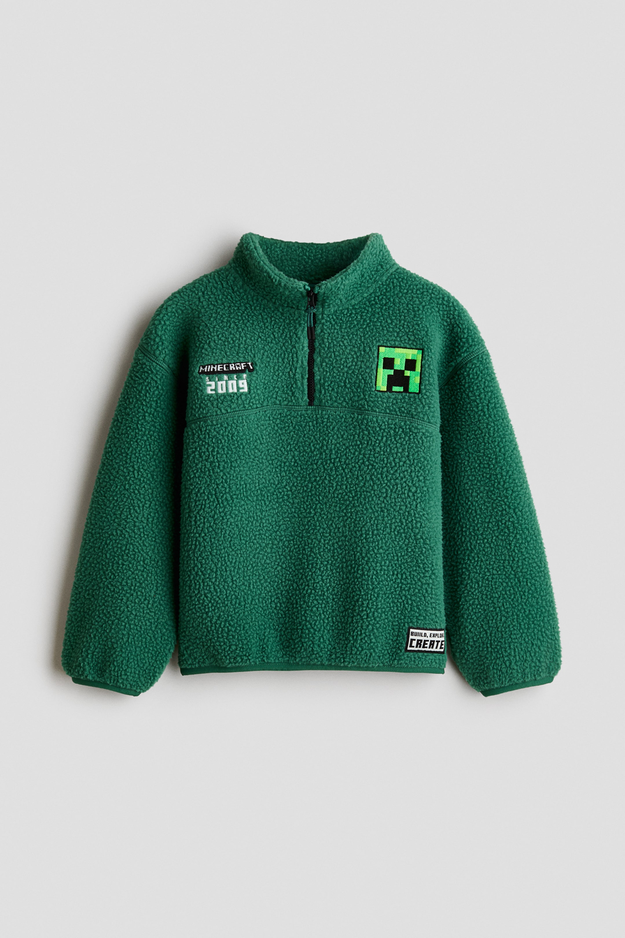View larger image: A green teddy fleece sweater is shown from the front, featuring a stand collar, a black half-zip closure, dropped shoulders, and long sleeves. The sweater features Minecraft-themed designs, including a Creeper face patch on the chest, embroidered text on the right side, and matching green jersey binding along the cuffs and hem.