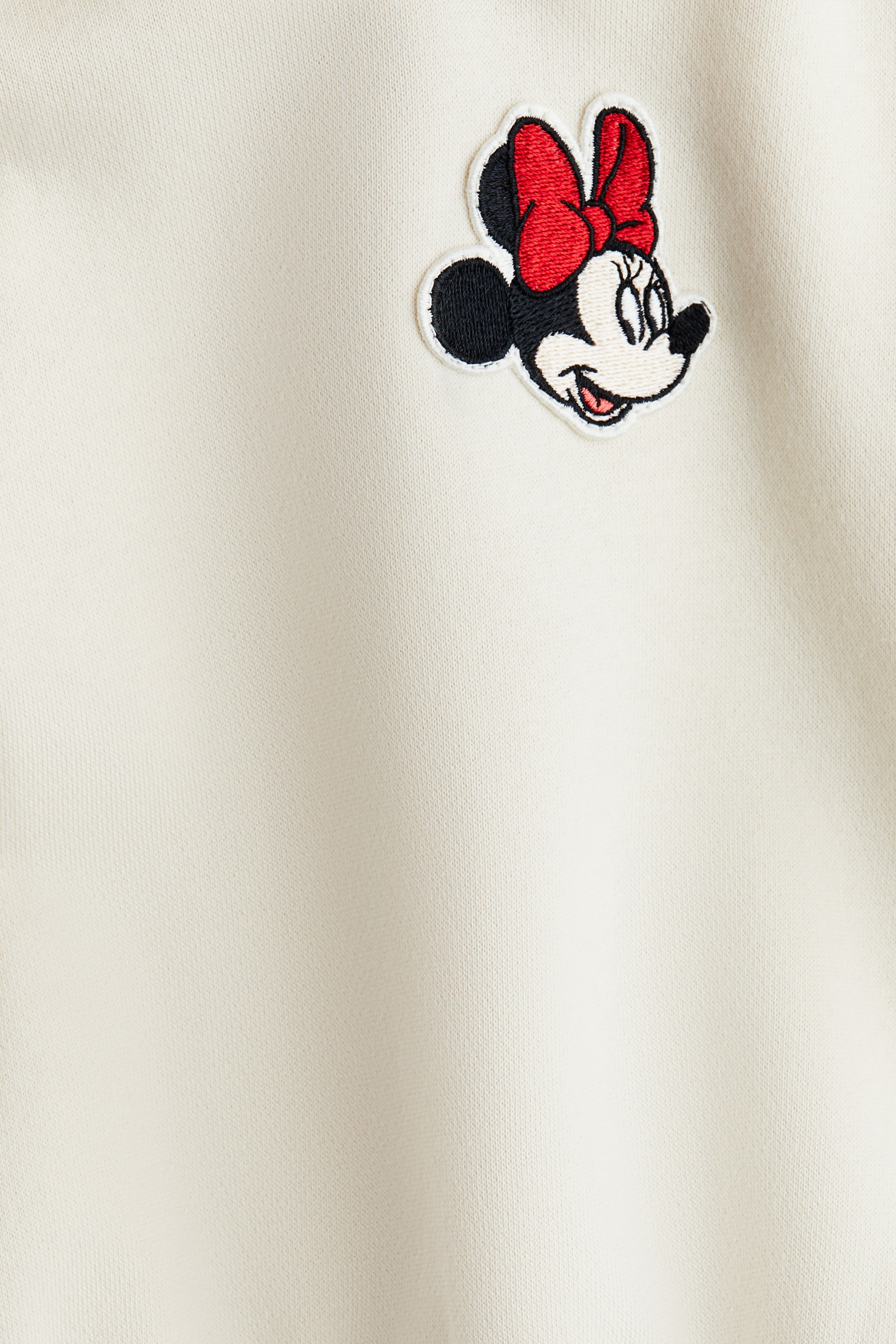 Zobraziť väčší obrázok: Krémová látka obsahuje vyšívanú nášivku hlavy Minnie Mouse, ktorá je detailne spracovaná s čiernymi ušami, bielou tvárou a červenou mašľou.