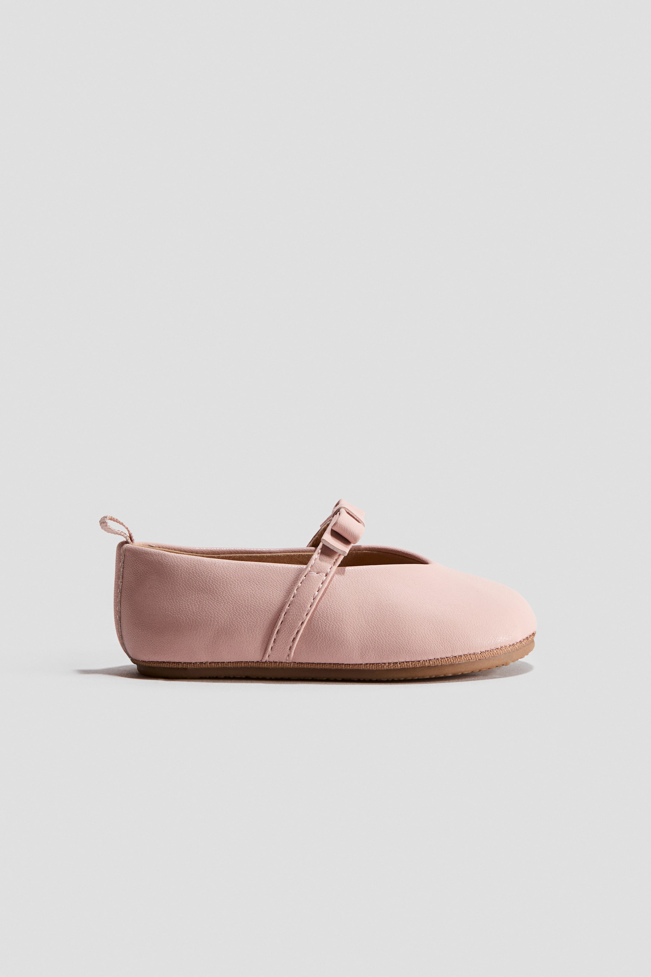 Grösseres Bild ansehen: Ein hellrosa Ballettschuh mit runder Spitze, einem Riemen mit Schleifendetail über dem Fuß, einer kleinen Schlaufe am Rücken und einer geriffelten Sohle.