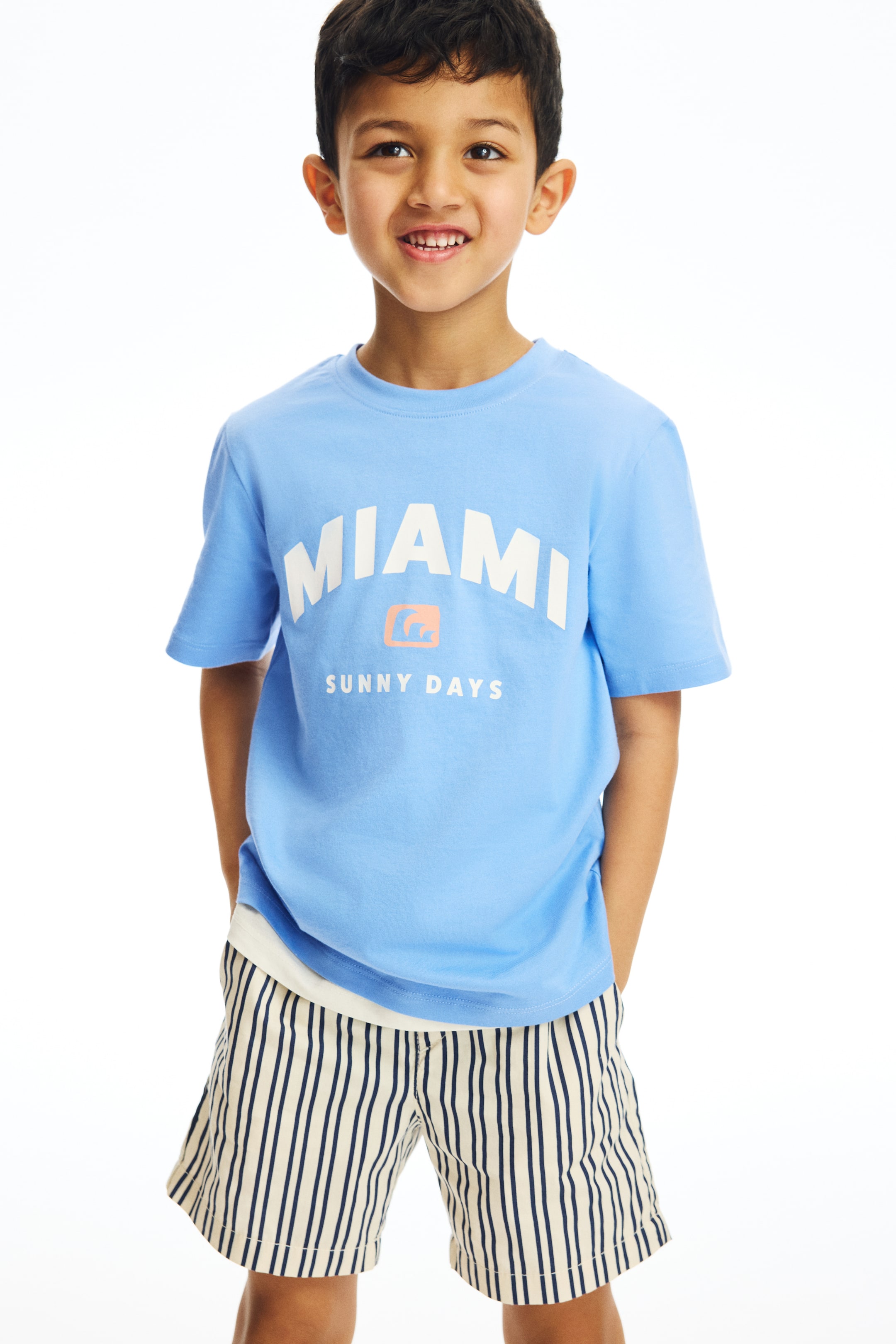 View larger image: A light blue short-sleeved t-shirt with a crew neck and a "MIAMI" graphic in arched white letters, an orange icon, and "SUNNY DAYS" below, is being presented front-facing. It is being worn over a white undershirt and paired with cream shorts featuring narrow vertical navy blue stripes, with hands resting in the pockets.