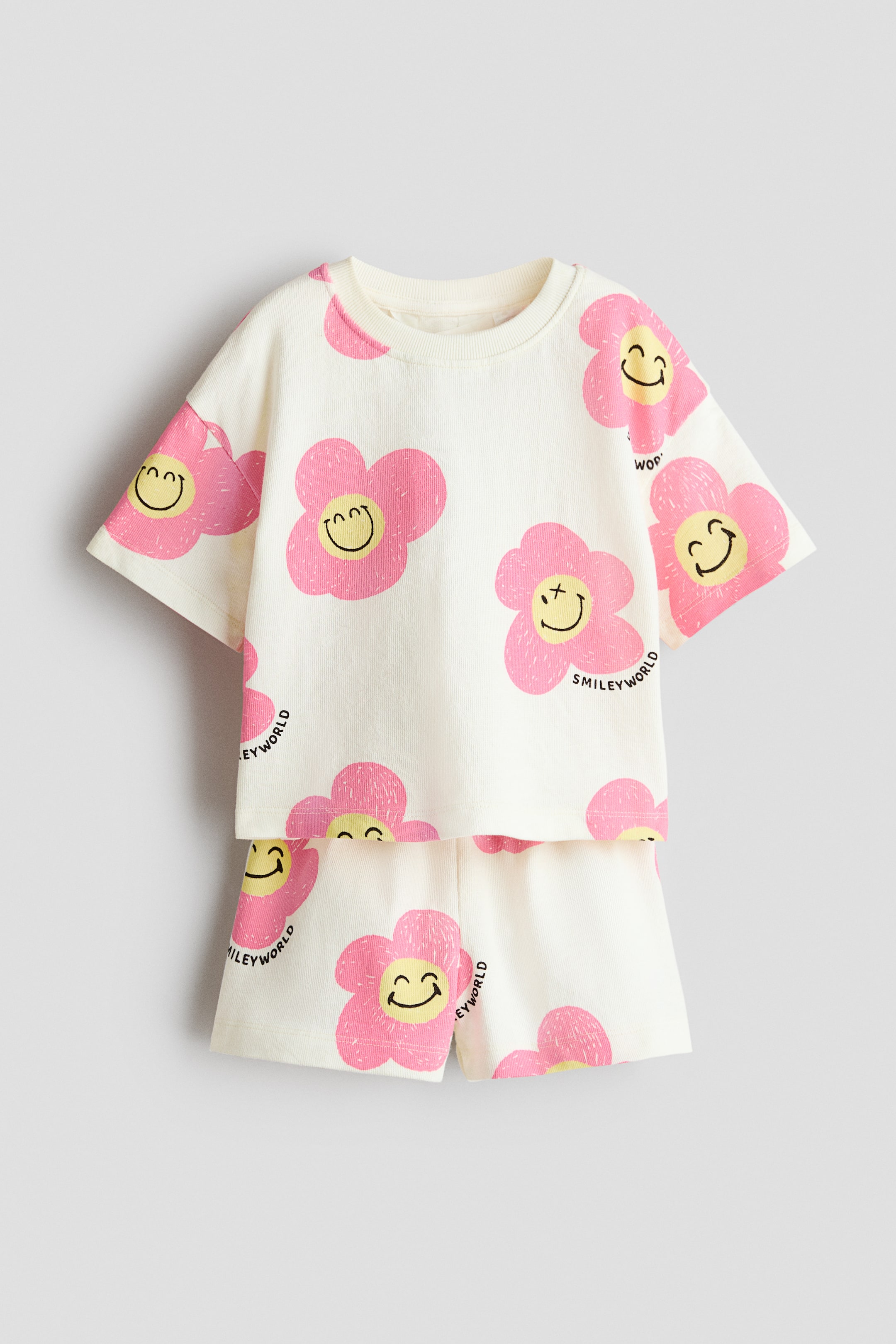 Agrandir l'image: Un ensemble composé d'un T-shirt blanc cassé et d'un short assorti est présenté à plat, face avant. Le T-shirt arbore un imprimé intégral de fleurs roses avec des visages souriants jaunes, un col rond avec bord-côtes et des épaules tombantes, tandis que le short présente une taille élastiquée avec le même imprimé.