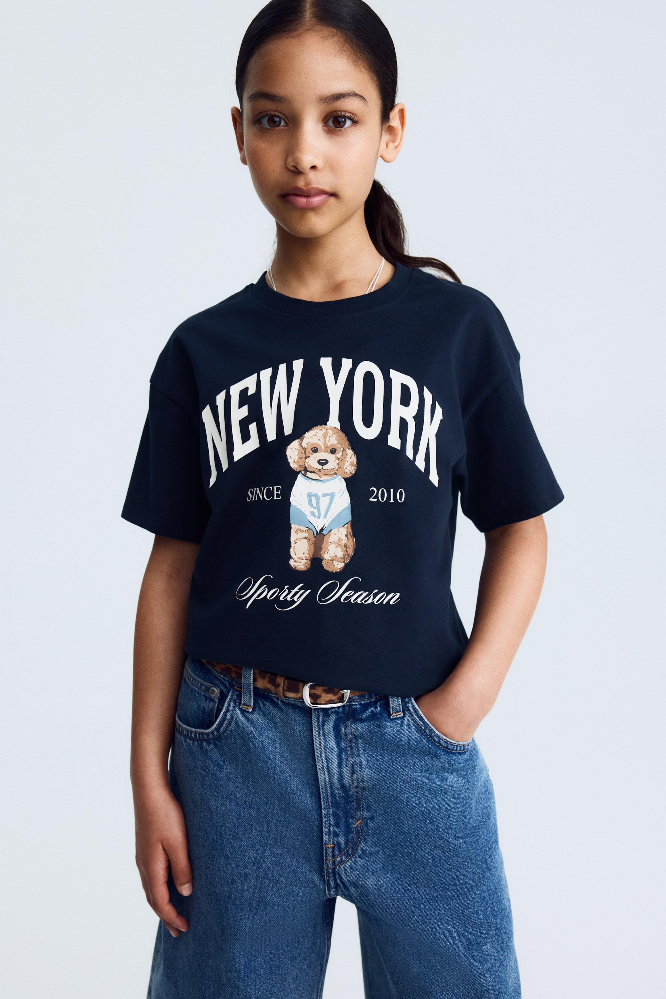 View larger image: A dark blue cotton jersey T-shirt is showcased from the front, featuring a round, rib-trimmed neckline and dropped shoulders. The front of the T-shirt displays white "NEW YORK" text, a brown poodle wearing a light blue shirt, and additional white text including "SINCE 2010" and "Sporty Season."