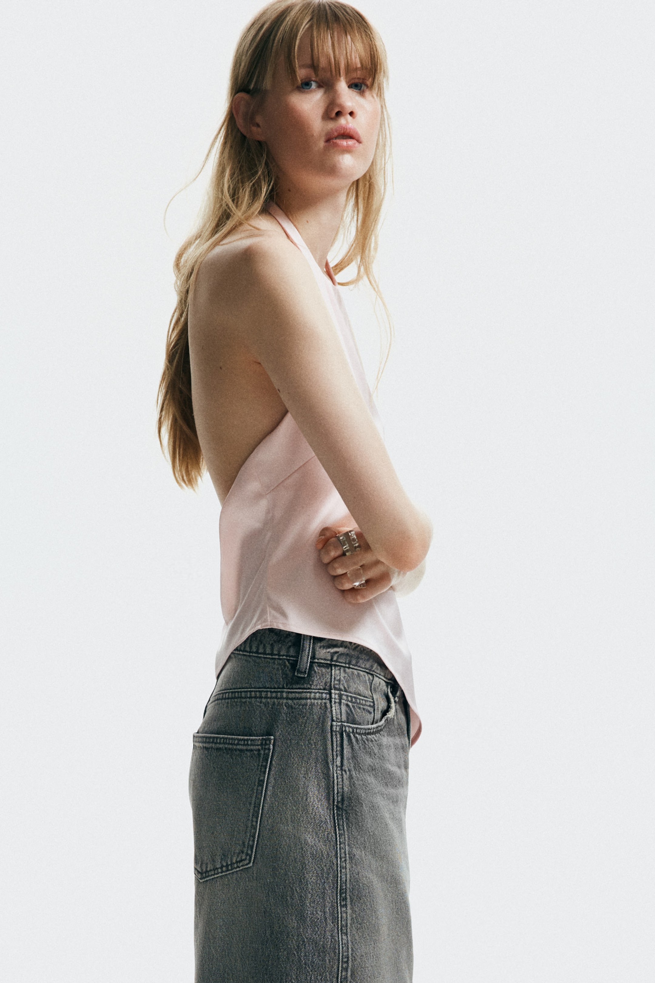 Größeres Bild anzeigen: Ein hellrosa Neckholder-Top mit einem fließenden, rückenfreien Design wird aus einem seitlichen Winkel getragen. Das Top ist in einen grauen Denim-Midirock gesteckt, der sichtbare Vorder- und Gesäßtaschen sowie Gürtelschlaufen aufweist.