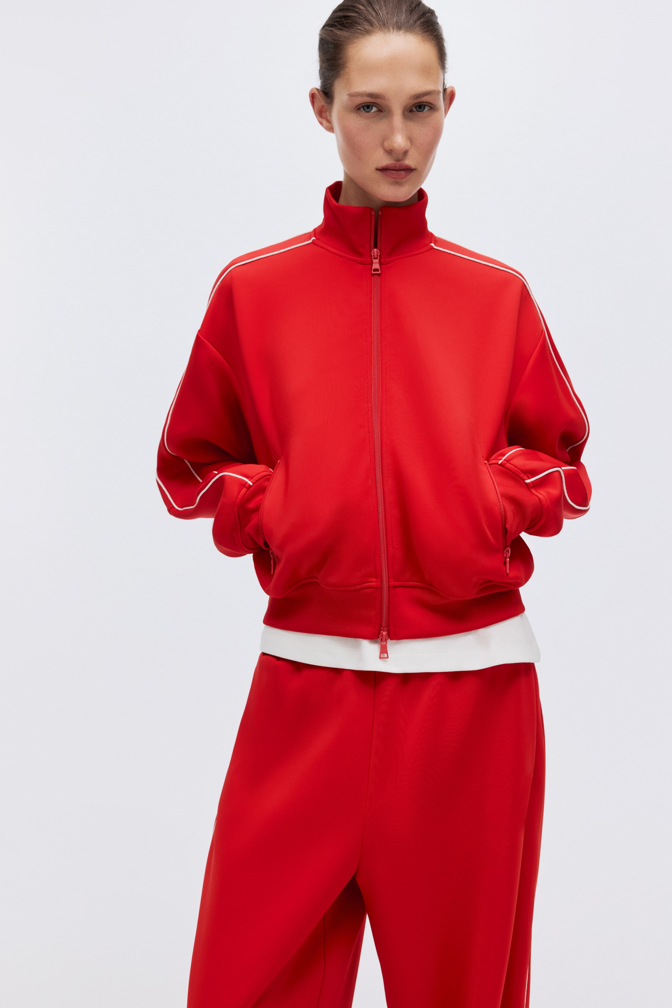 View larger image: A red tracksuit jacket features a full-zip closure, a high stand-up collar and white piping along the raglan sleeves and collar. The jacket offers a relaxed fit with ribbed cuffs and hem, layered over a white T-shirt, while matching loose-fitting red pants complete the ensemble.