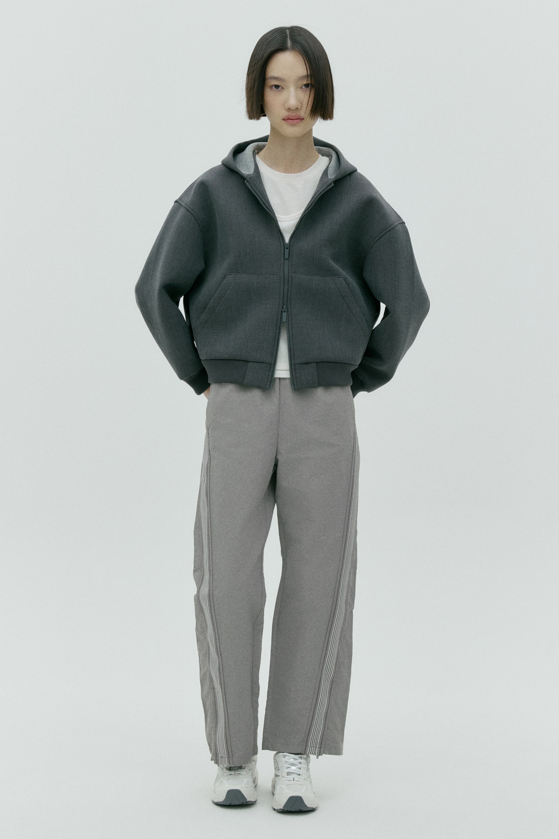 拡大画像を表示: ダークグレーのフード付きジップアップジャケット（オーバーサイズフィット）がホワイトTシャツの上に重ねられており、フロントにはフルジッパー、下部前面には大きく斜めに配されたポケットが施されています。ライトグレーのワイドパンツは縦ストライプのサイドパネル入りで、ホワイト×グレーのスニーカーの上にかかる丈がコーディネートを引き締めています。