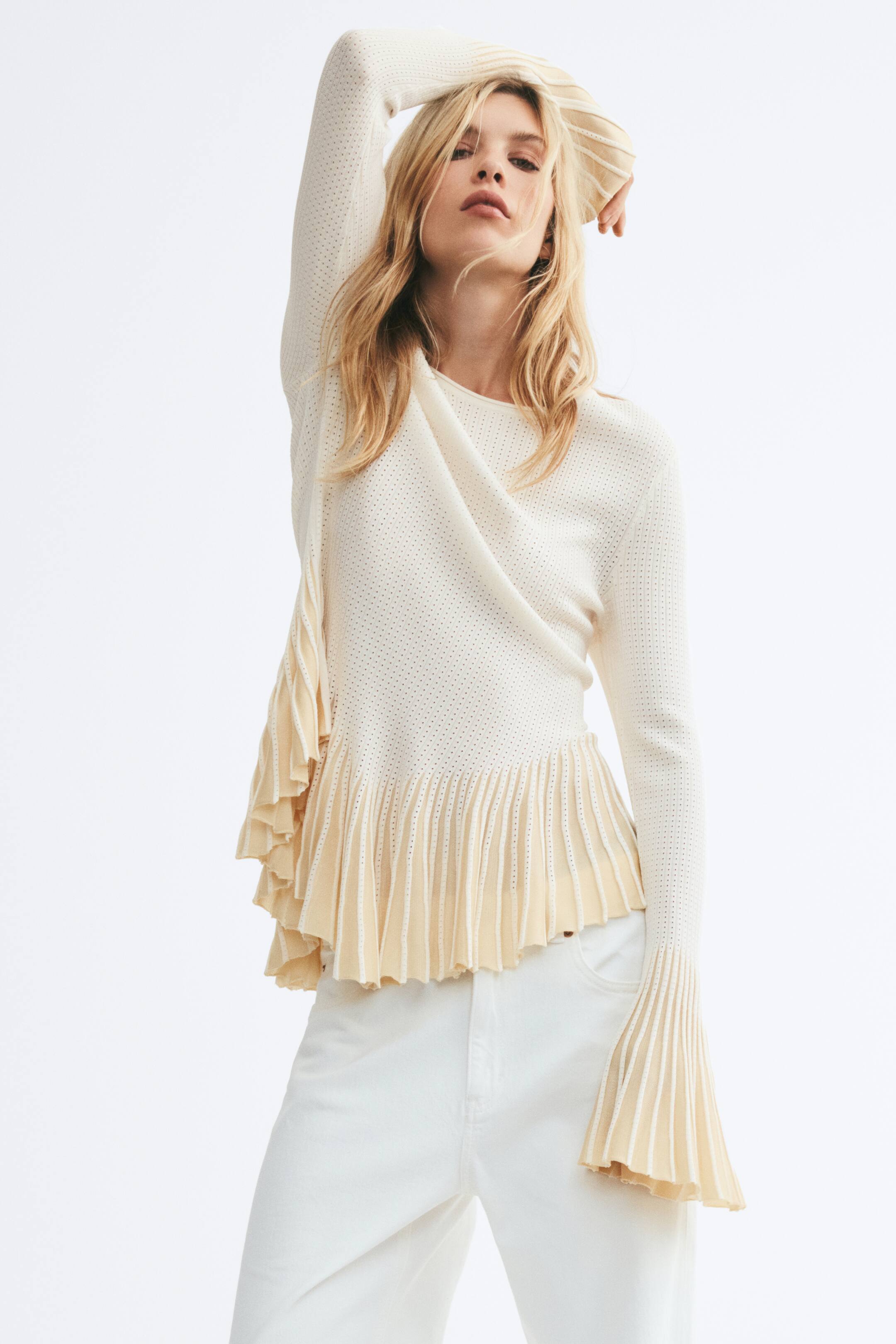 View larger image: The model is wearing a cream pointelle-knit top with a round neckline, long sleeves with pleated, flounced cuffs, and a matching flounced hem. She is styling it with white pants and posing with one arm raised.