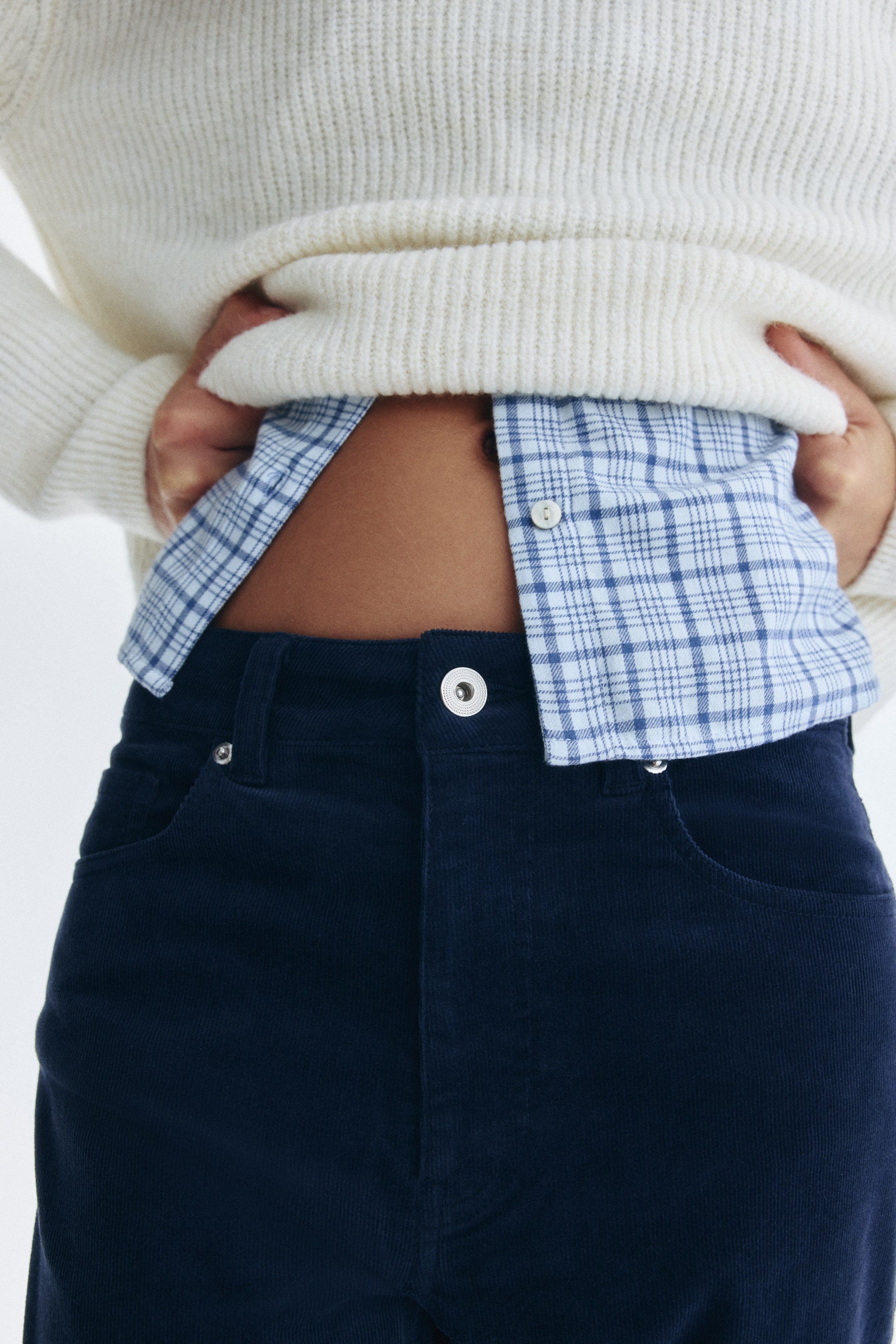 Voir une image plus grande: Un pantalon en velours côtelé bleu foncé est porté, avec une fermeture à bouton argenté, des passants de ceinture et des poches avant. Le mannequin soulève un pull en tricot côtelé couleur crème, dévoilant la ceinture du pantalon et une chemise à carreaux bleu clair rentrée dedans.
