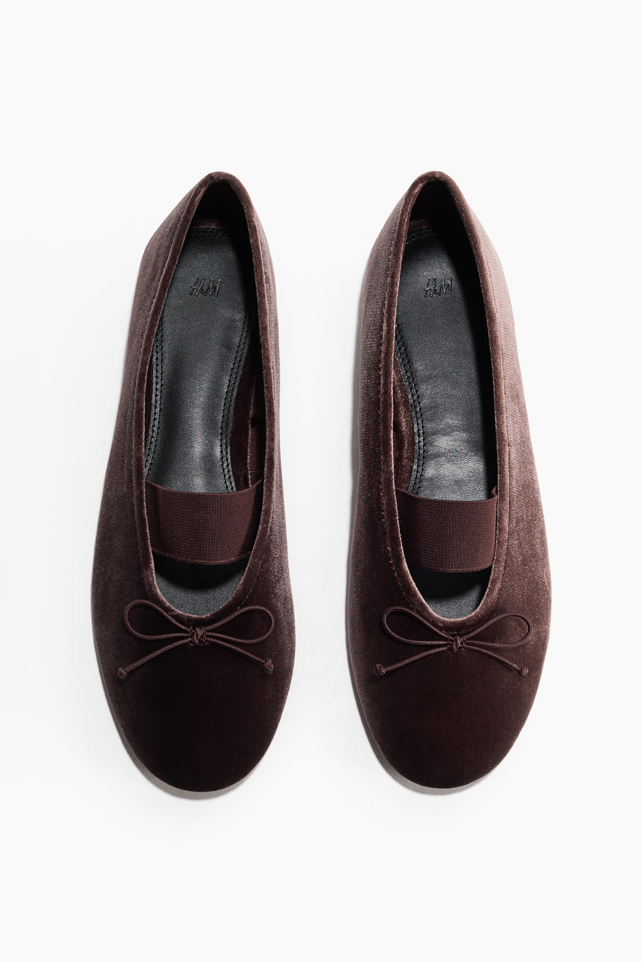 View larger image: A pair of dark brown velvet ballet flats is being showcased, highlighting the front toe and instep. Each flat is adorned with a small bow tie on the toe and an elastic strap stretching across the instep. The black insole displays the H&M logo.