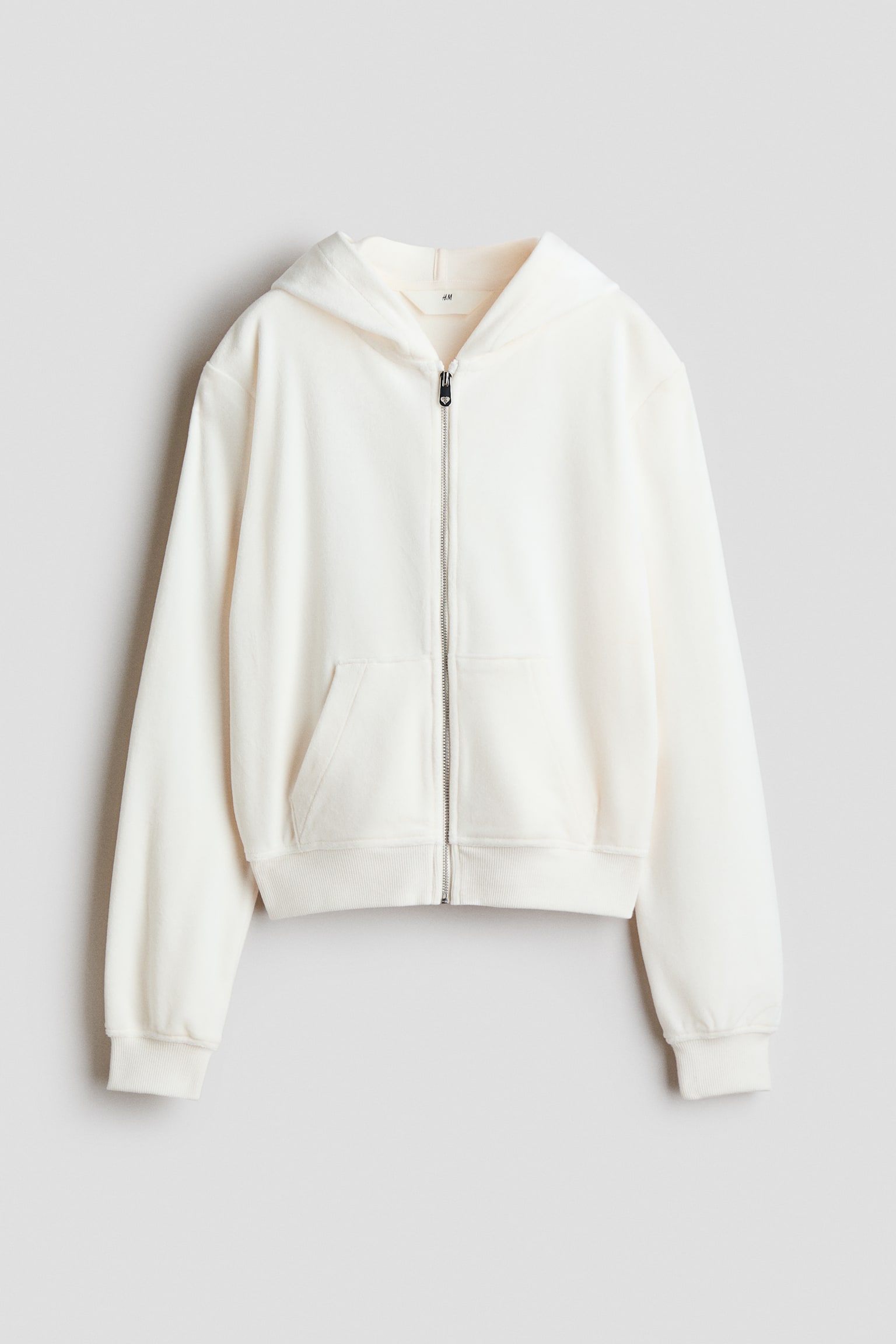 Un hoodie zippÃ© en velours blanc cassÃ© est prÃ©sentÃ© de face, avec une capuche doublÃ©e en jersey, des Ã©paules tombantes et une fermeture Ã©clair frontale argentÃ©e. Le hoodie prÃ©sente des poches kangourou fendues Ã  lâavant, ainsi que de larges bords-cÃ´tes aux poignets et Ã  lâourlet.