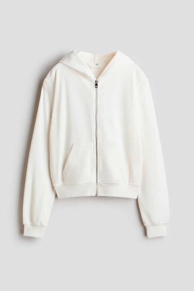 An off-white velour zip-up hoodie is shown from the front, featuring a jersey-lined hood, dropped shoulders, and a silver front zip. The hoodie features split front kangaroo pockets, along with wide ribbed trims at the cuffs and hem. - 1
