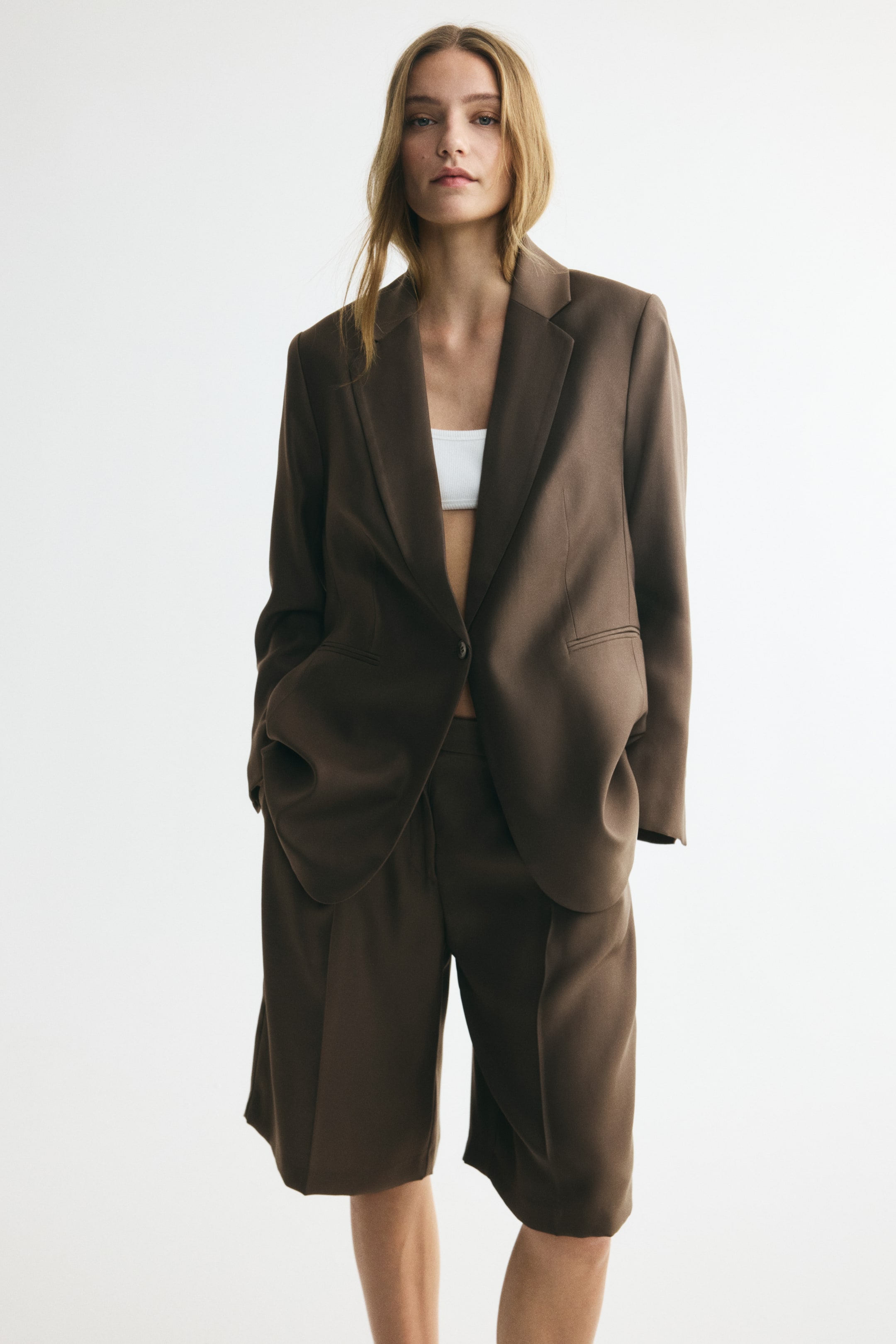 View larger image: A long, dark brown single-breasted blazer is presented front-on, featuring a relaxed fit, notch lapels, and jetted front pockets. It showcases a single-button fastening and is worn open over a white bralette.