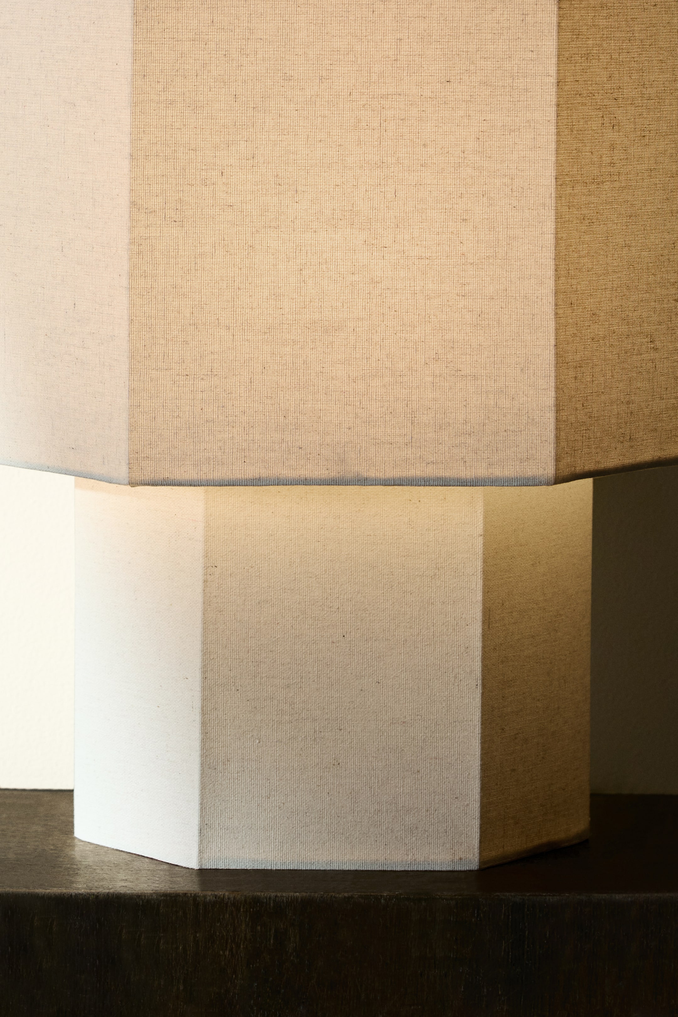 View larger image: A multi-faceted lampshade, crafted from light-coloured, textured fabric, is casting light from within. It rests on a matching multi-faceted lamp base, also fashioned from textured fabric, with its vertical panels displaying varying shades of light and shadow.