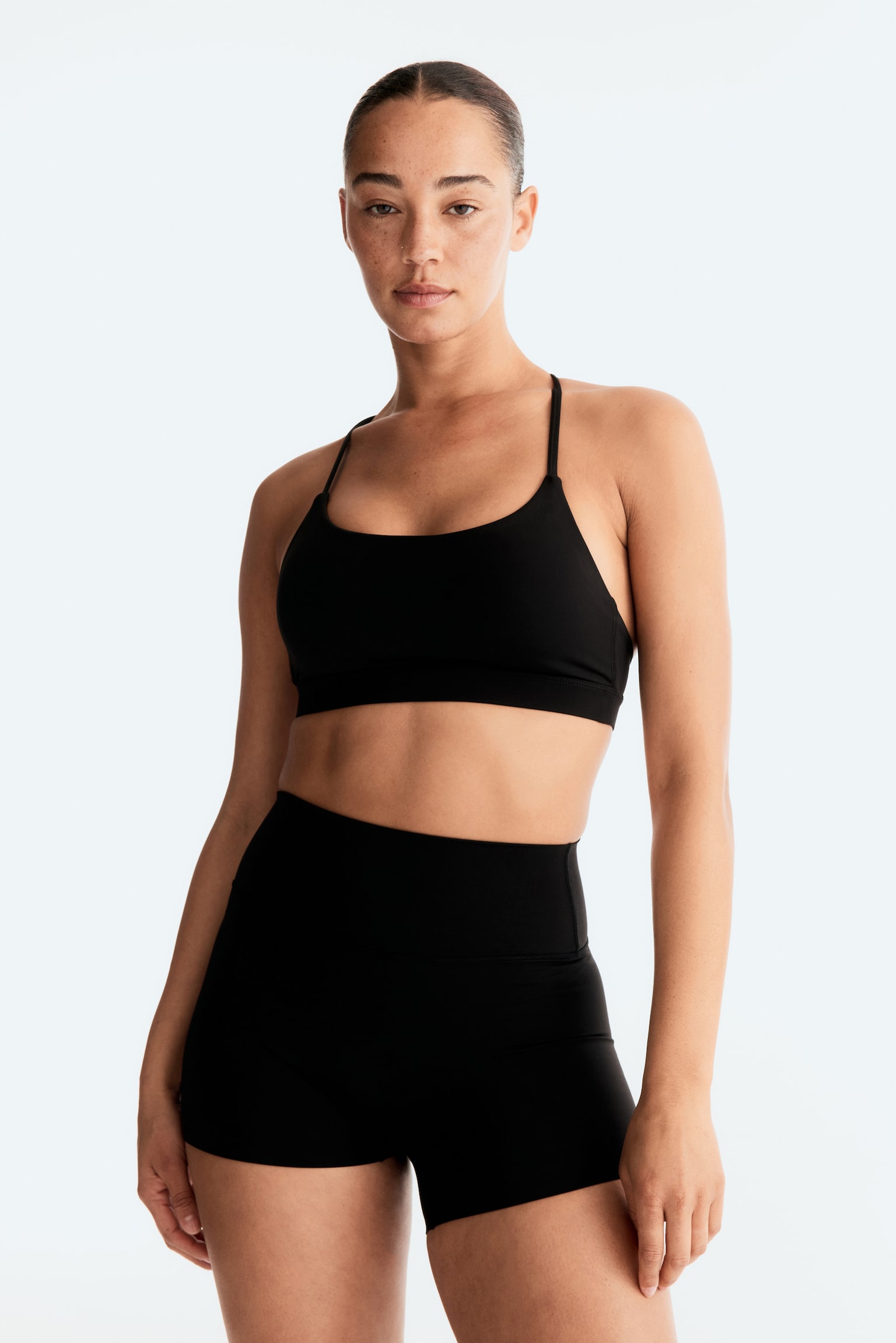 A black sports bra and matching black shorts are being displayed front-facing. The sports bra features a scoop neck, thin shoulder straps, and a wide elasticized band under the bust. The high-waisted shorts have a wide, flat waistband and a short, form-fitting length. - 1
