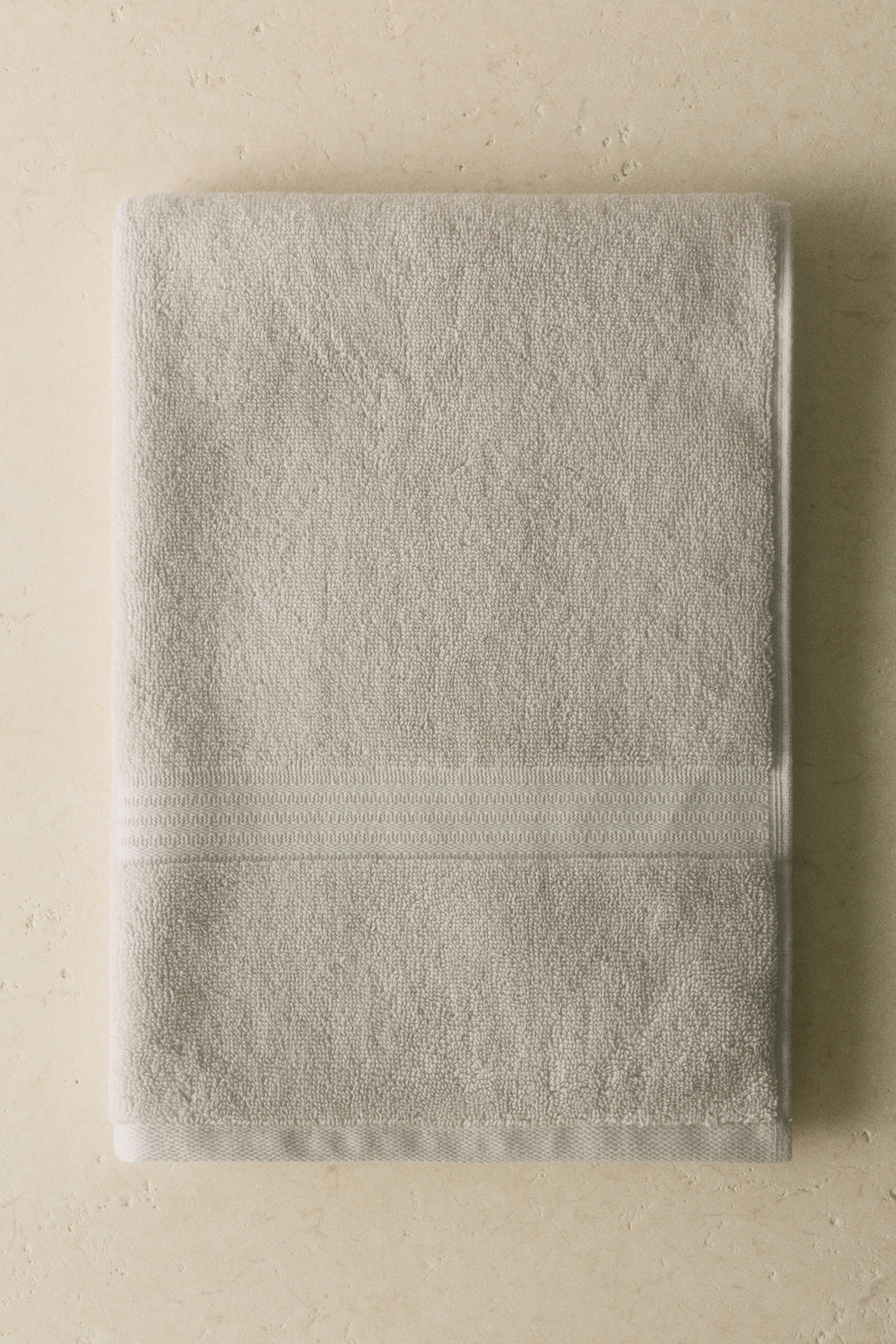 Une serviette de toilette gris clair est suspendue, affichant sa texture en éponge de coton avec un éclat subtil, une bande de garniture tissée à une extrémité courte, et une boucle de suspension visible à l'autre.