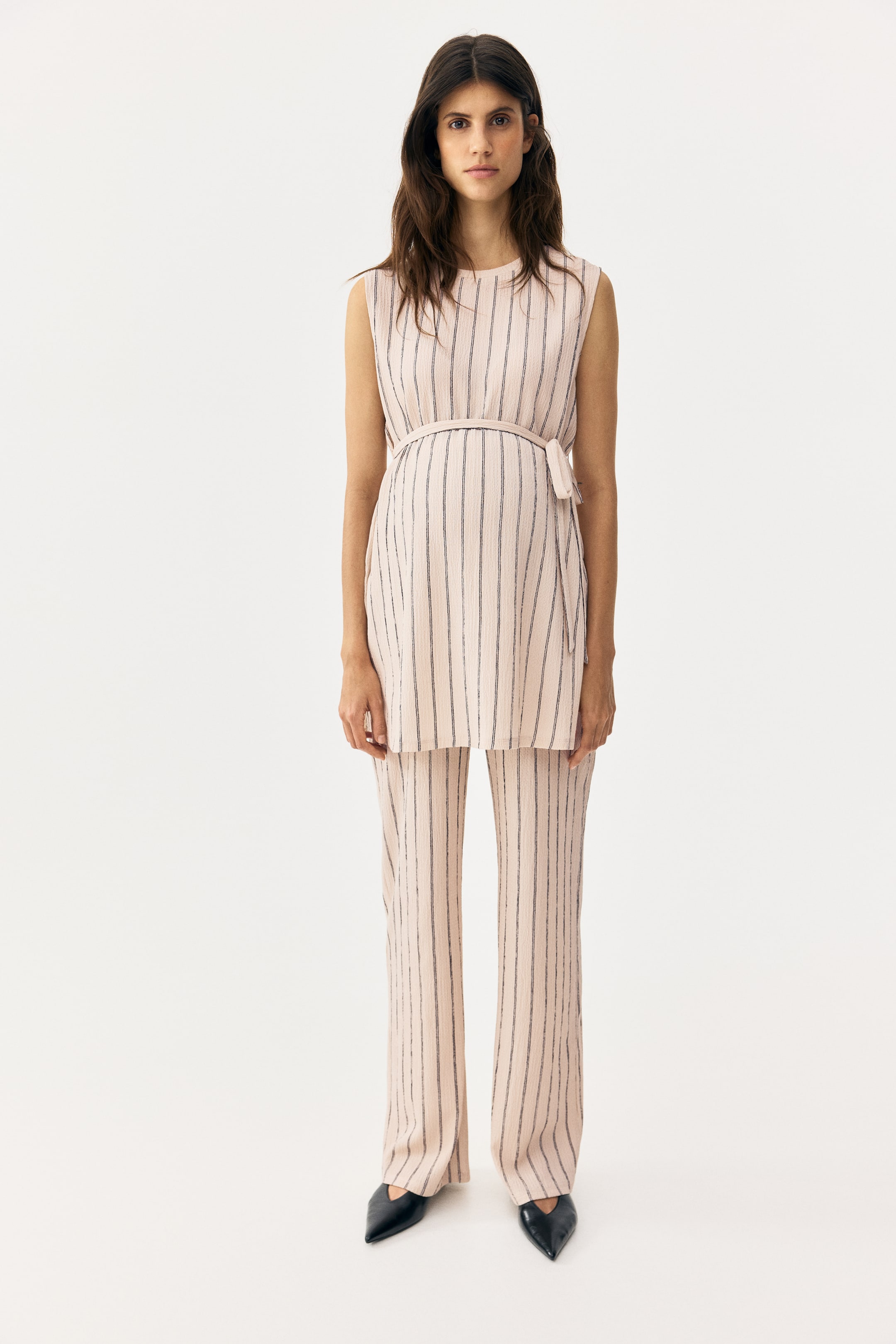 View larger image: A light pink two-piece outfit features a sleeveless tunic top with a round neckline and wide-leg pants, both adorned with thin dark vertical stripes. The tunic cinches at the waist with a matching fabric belt, tied into a small bow on the right side, and the entire outfit is shown from the front.