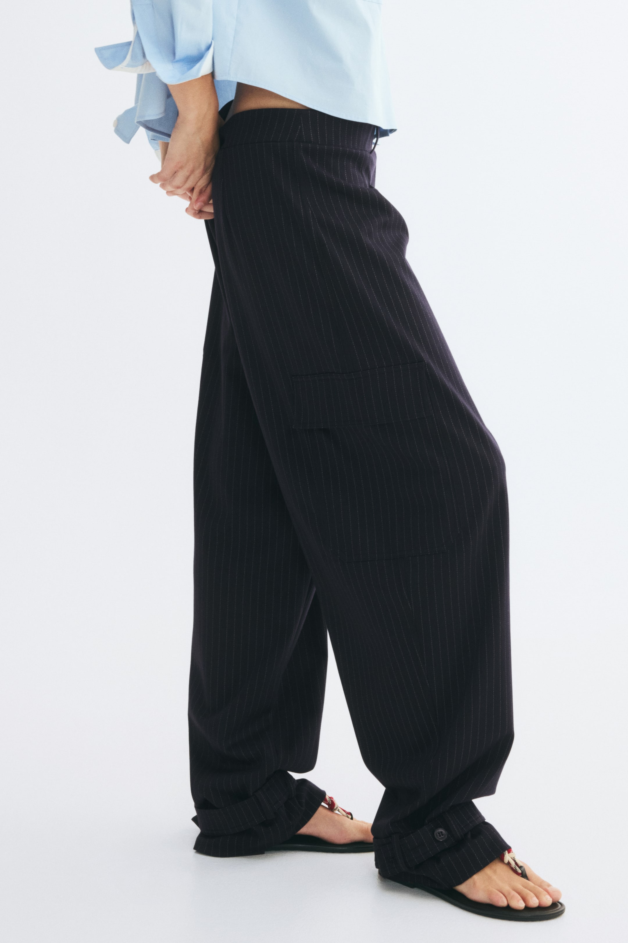 View larger image: Loose-fit cargo trousers in dark pinstriped jacquard jersey are facing left, showcasing a wide-leg silhouette and a side flap pocket. The trousers sit at the waist and taper at the ankles with adjustable button-strap hems.