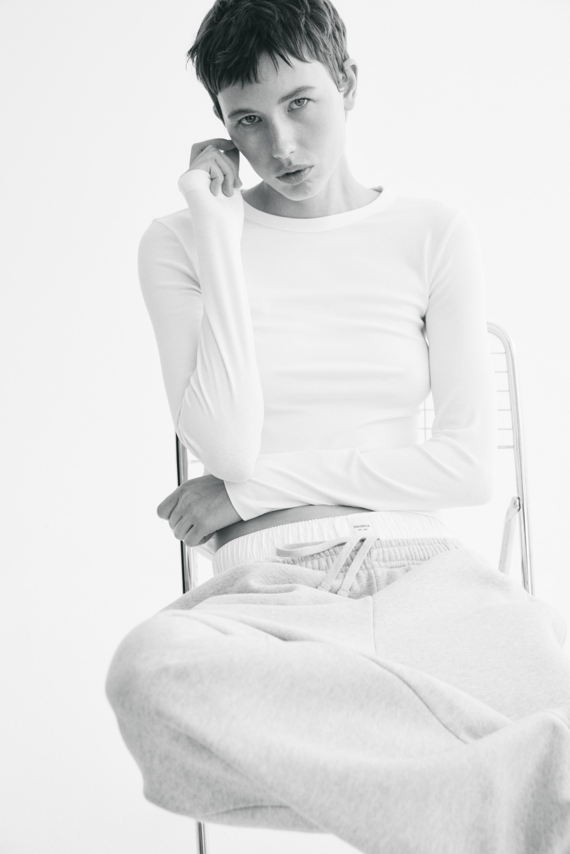 View larger image: The model is wearing a white, fitted, ribbed jersey top featuring a round neckline and long sleeves. She is seated, with one hand resting on her face, and the sleeve of the top elegantly drapes over her forearm. Complementing the look, she is also wearing light grey sweatpants.