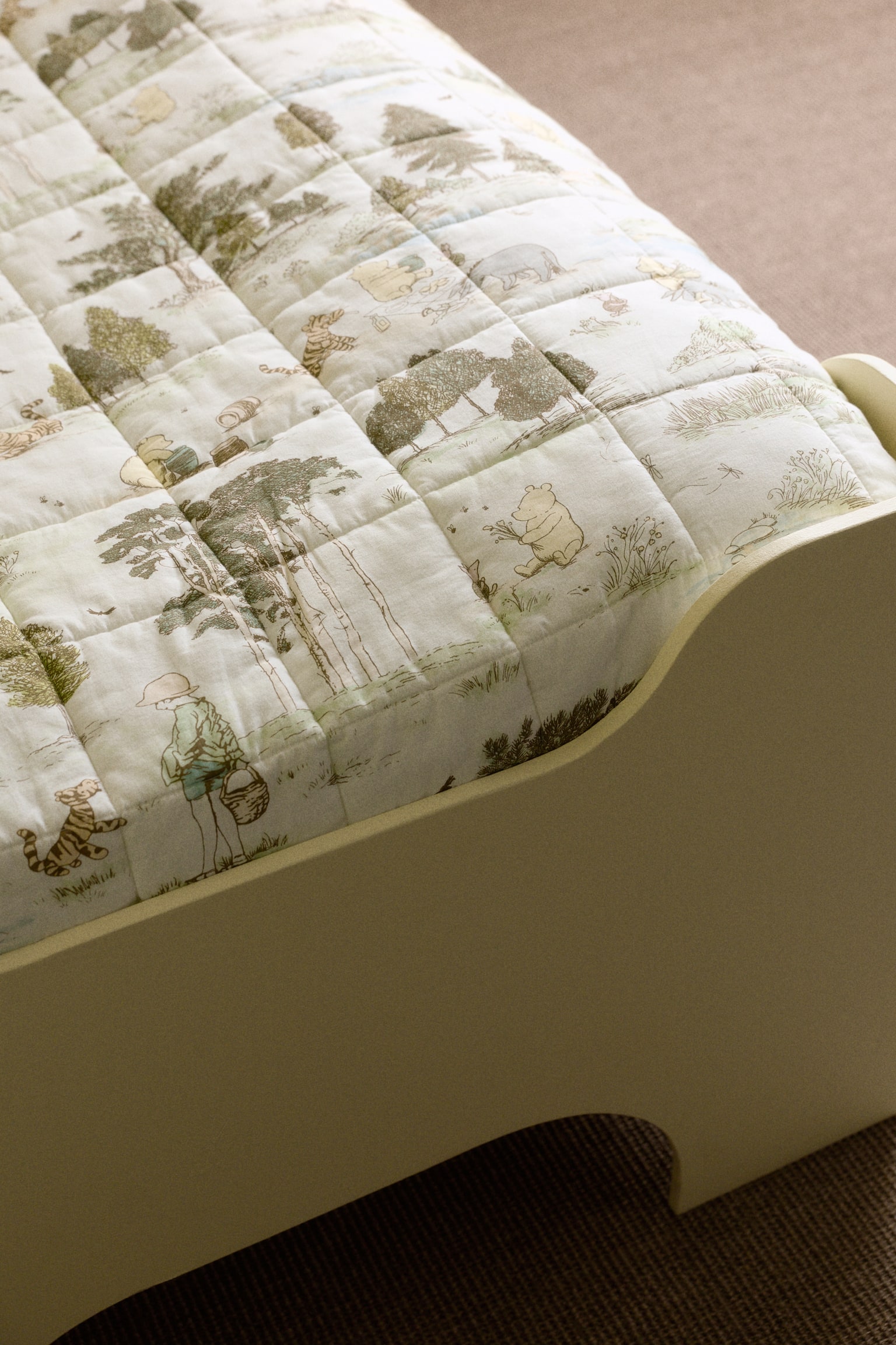 A quilted blanket is lying flat on a bed, showcasing a grid pattern and an illustrated scenic print featuring forest elements and characters like Winnie the Pooh, Tigger, and Christopher Robin. It rests on a light-coloured bed frame with a curved side rail, viewed from a slight angle. - 1